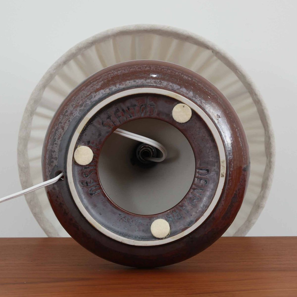 デンマークのDesireeによる陶磁器製のテーブルランプ。深みのある赤褐色を基調に、黒やグレーが複雑に溶け合う釉薬の表情が印象的な一台です。裾に向かって大きく広がる丸みを帯びたフォルムと、すっきりと伸びる首元のバランスが美しく、陶磁器ならではの重厚感と優雅さを兼ね備えています。細かなプリーツを施したリネンシェードは、点灯すると柔らかな光を透過し、温かみのある落ち着いた雰囲気を演出します。消灯時にもオブジェのような存在感があり、リビングのサイドボードや寝室のベッドサイドなど、さまざまな空間におすすめです。釉薬の豊かな色彩と質感に、北欧ビンテージ陶器ならではの魅力が感じられます。