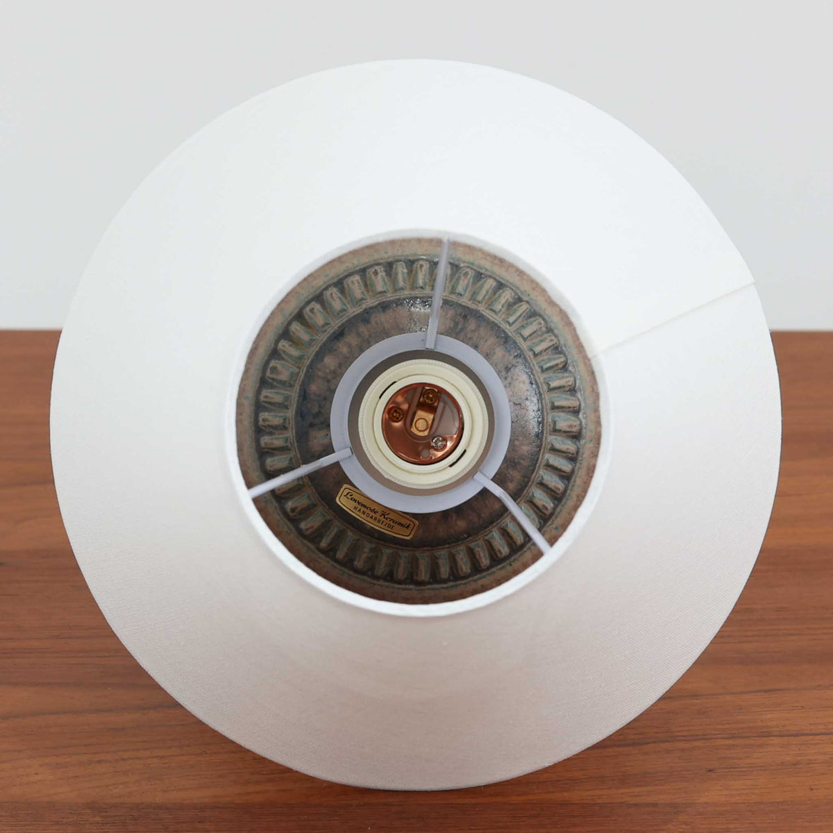 デンマークのLovemose Keramikによる陶磁器製のテーブルランプ。ふっくらと丸みを帯びたボディから細く伸びる首元へと続く、優美でバランスの取れたフォルムが印象的な一台です。ブラウンやグレーが複雑に混ざり合う釉薬には、流れるような濃淡や細かな斑点模様が現れ、陶磁器ならではの豊かな表情を楽しめます。首元と胴体の境目には連続した彫り模様が施され、素朴な風合いの中に職人の丁寧な手仕事が感じられます。シンプルなリネンシェードを組み合わせることで、点灯時には柔らかな光が広がり、落ち着いた空間を演出します。リビングのサイドボードや寝室のベッドサイドなどにおすすめの、北欧ビンテージらしい温もりと存在感を備えた照明です。