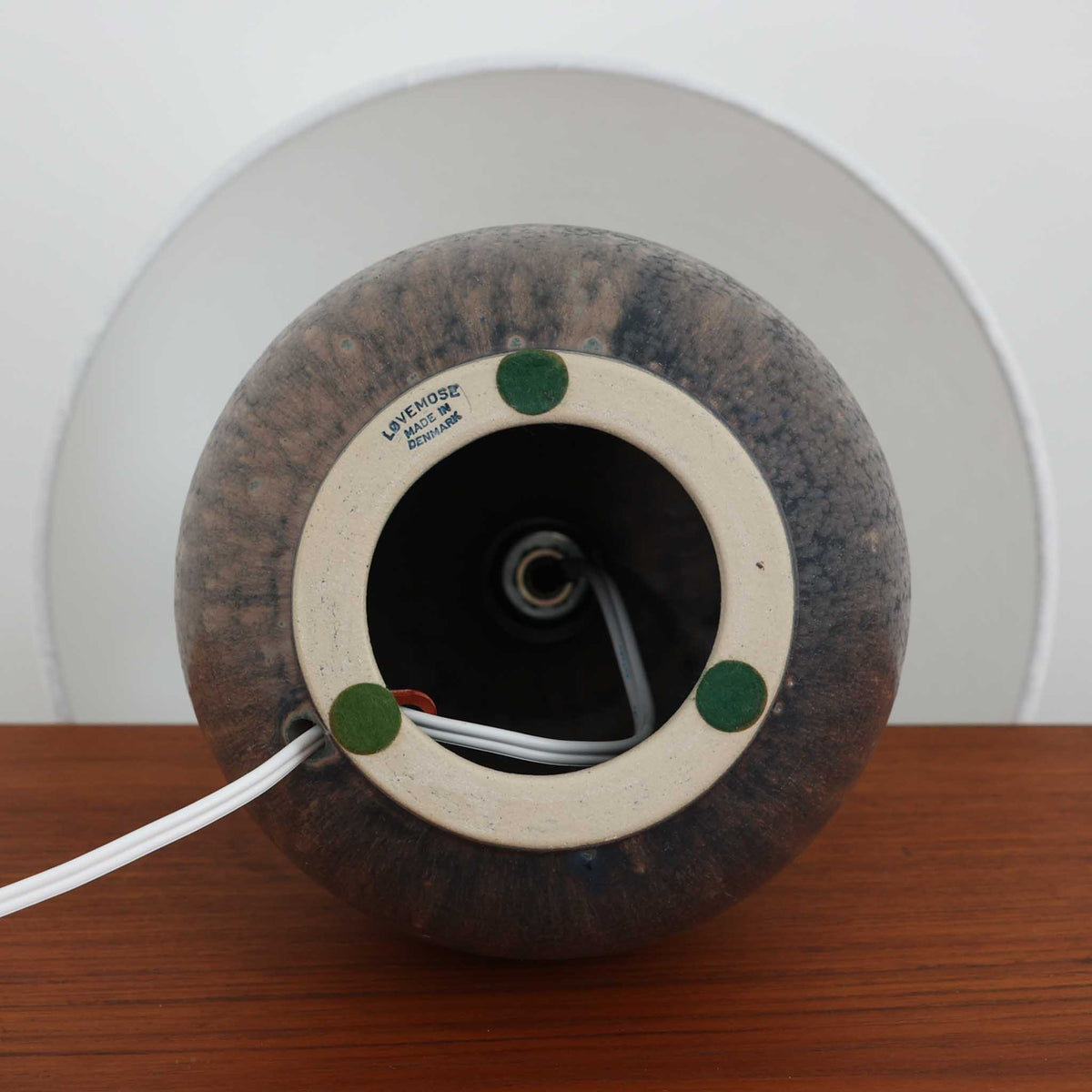 デンマークのLovemose Keramikによる陶磁器製のテーブルランプ。ふっくらと丸みを帯びたボディから細く伸びる首元へと続く、優美でバランスの取れたフォルムが印象的な一台です。ブラウンやグレーが複雑に混ざり合う釉薬には、流れるような濃淡や細かな斑点模様が現れ、陶磁器ならではの豊かな表情を楽しめます。首元と胴体の境目には連続した彫り模様が施され、素朴な風合いの中に職人の丁寧な手仕事が感じられます。シンプルなリネンシェードを組み合わせることで、点灯時には柔らかな光が広がり、落ち着いた空間を演出します。リビングのサイドボードや寝室のベッドサイドなどにおすすめの、北欧ビンテージらしい温もりと存在感を備えた照明です。