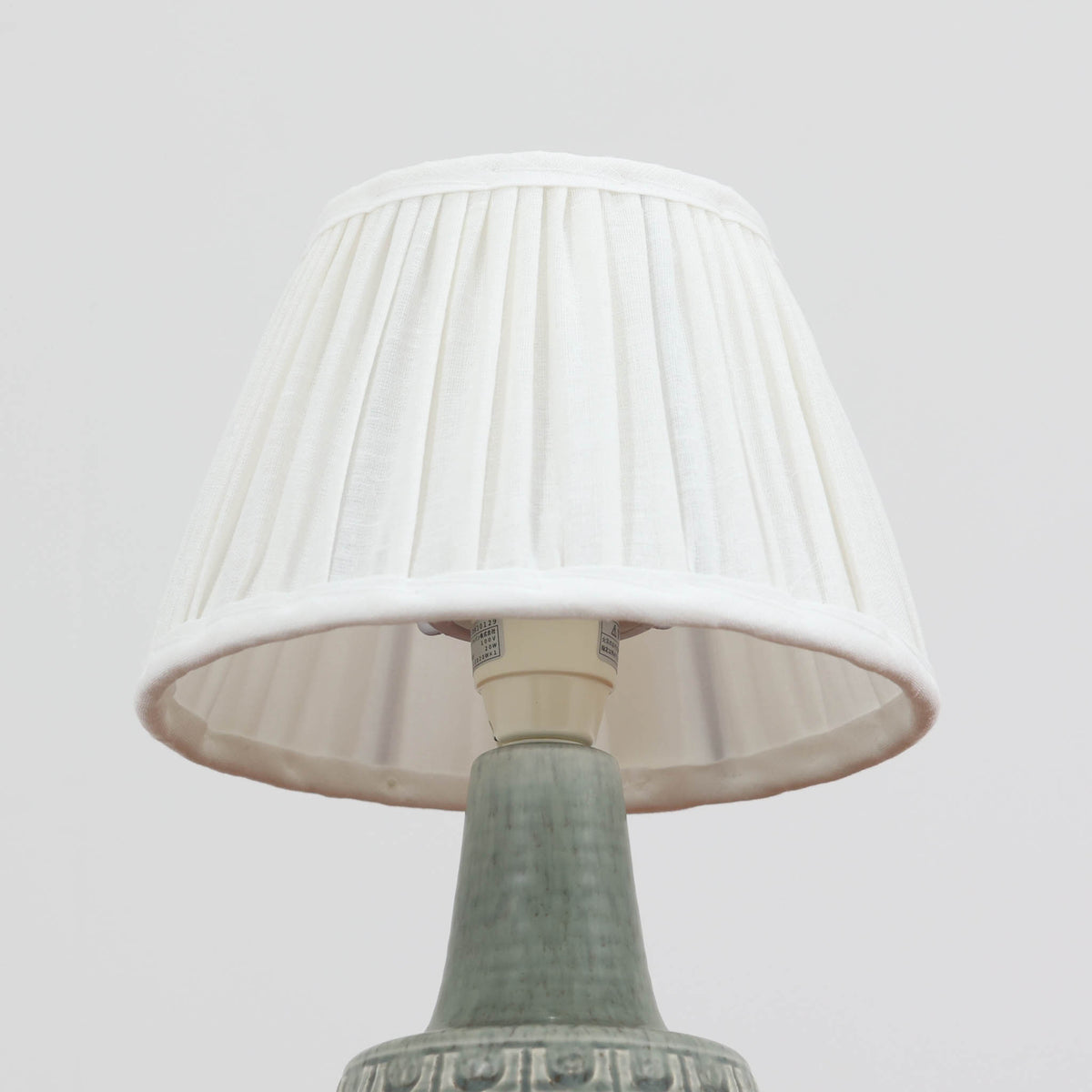 デンマークの陶磁器メーカーSoholm Keramik（スーホルム）による、Einar Johansen（アイナー・ヨハンセン）デザインのテーブルランプ。落ち着いたグリーングレーの釉薬が美しい陶磁器製のベースは、ふっくらとした下部からすっきりと伸びる首元へと続く、優雅なフォルムが特徴です。胴体には幾何学的なレリーフ模様が帯状に施され、釉薬の繊細な濃淡と相まって豊かな立体感を生み出しています。細かなプリーツを施したリネンシェードは、点灯すると柔らかな光を透過し、温かみのある落ち着いた雰囲気を演出します。北欧ビンテージ陶器ならではの味わい深い質感と造形美を楽しめる一台で、リビングのサイドボードや寝室のベッドサイドなどにおすすめです。