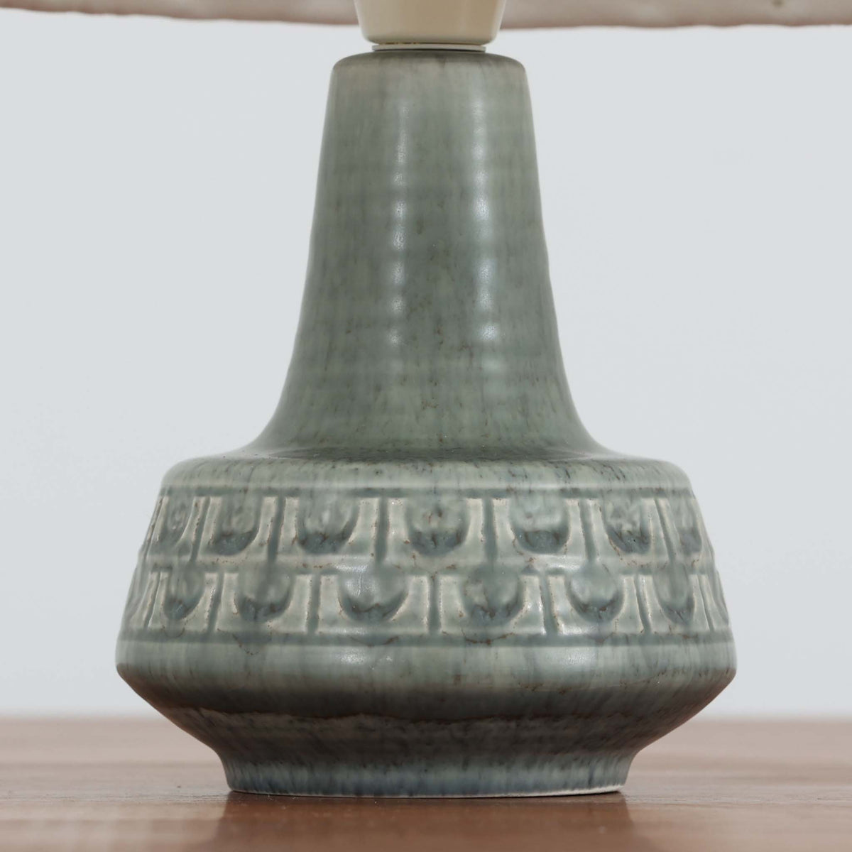デンマークの陶磁器メーカーSoholm Keramik（スーホルム）による、Einar Johansen（アイナー・ヨハンセン）デザインのテーブルランプ。落ち着いたグリーングレーの釉薬が美しい陶磁器製のベースは、ふっくらとした下部からすっきりと伸びる首元へと続く、優雅なフォルムが特徴です。胴体には幾何学的なレリーフ模様が帯状に施され、釉薬の繊細な濃淡と相まって豊かな立体感を生み出しています。細かなプリーツを施したリネンシェードは、点灯すると柔らかな光を透過し、温かみのある落ち着いた雰囲気を演出します。北欧ビンテージ陶器ならではの味わい深い質感と造形美を楽しめる一台で、リビングのサイドボードや寝室のベッドサイドなどにおすすめです。