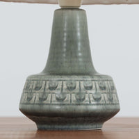 デンマークの陶磁器メーカーSoholm Keramik（スーホルム）による、Einar Johansen（アイナー・ヨハンセン）デザインのテーブルランプ。落ち着いたグリーングレーの釉薬が美しい陶磁器製のベースは、ふっくらとした下部からすっきりと伸びる首元へと続く、優雅なフォルムが特徴です。胴体には幾何学的なレリーフ模様が帯状に施され、釉薬の繊細な濃淡と相まって豊かな立体感を生み出しています。細かなプリーツを施したリネンシェードは、点灯すると柔らかな光を透過し、温かみのある落ち着いた雰囲気を演出します。北欧ビンテージ陶器ならではの味わい深い質感と造形美を楽しめる一台で、リビングのサイドボードや寝室のベッドサイドなどにおすすめです。