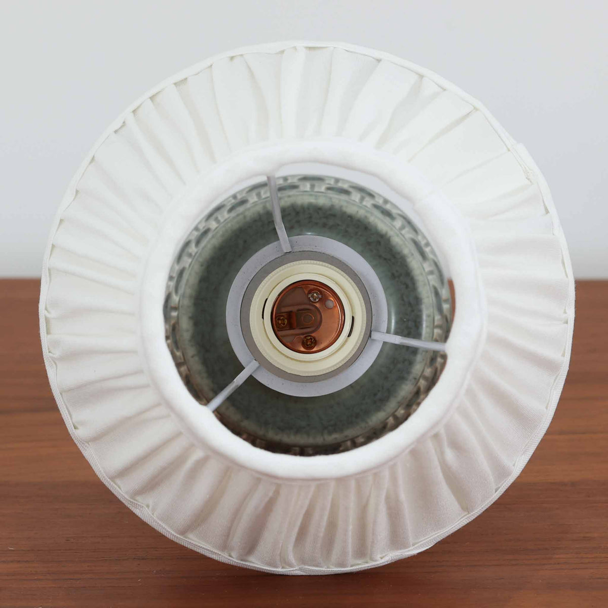 デンマークの陶磁器メーカーSoholm Keramik（スーホルム）による、Einar Johansen（アイナー・ヨハンセン）デザインのテーブルランプ。落ち着いたグリーングレーの釉薬が美しい陶磁器製のベースは、ふっくらとした下部からすっきりと伸びる首元へと続く、優雅なフォルムが特徴です。胴体には幾何学的なレリーフ模様が帯状に施され、釉薬の繊細な濃淡と相まって豊かな立体感を生み出しています。細かなプリーツを施したリネンシェードは、点灯すると柔らかな光を透過し、温かみのある落ち着いた雰囲気を演出します。北欧ビンテージ陶器ならではの味わい深い質感と造形美を楽しめる一台で、リビングのサイドボードや寝室のベッドサイドなどにおすすめです。