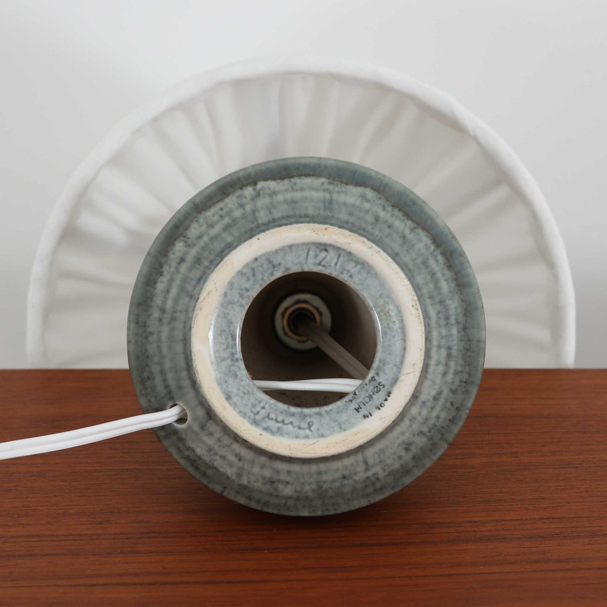 デンマークの陶磁器メーカーSoholm Keramik（スーホルム）による、Einar Johansen（アイナー・ヨハンセン）デザインのテーブルランプ。落ち着いたグリーングレーの釉薬が美しい陶磁器製のベースは、ふっくらとした下部からすっきりと伸びる首元へと続く、優雅なフォルムが特徴です。胴体には幾何学的なレリーフ模様が帯状に施され、釉薬の繊細な濃淡と相まって豊かな立体感を生み出しています。細かなプリーツを施したリネンシェードは、点灯すると柔らかな光を透過し、温かみのある落ち着いた雰囲気を演出します。北欧ビンテージ陶器ならではの味わい深い質感と造形美を楽しめる一台で、リビングのサイドボードや寝室のベッドサイドなどにおすすめです。