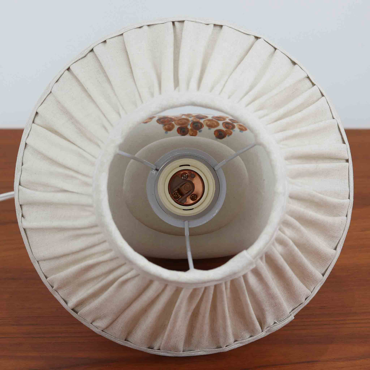 デンマークの陶磁器メーカーSoholm Keramik（スーホルム）による、Einar Paulsen（アイナー・ポールセン）デザインのテーブルランプ。柔らかなグレーベージュの陶磁器製ベースには、ブラウンやブルーを用いた植物を思わせる絵柄が描かれ、素朴で温かみのある雰囲気を醸し出しています。丸みを帯びたふっくらとしたフォルムと、首元に施された緩やかな曲線が美しく、手仕事ならではの豊かな表情が魅力です。細かなプリーツを施したリネンシェードは、点灯すると優しい光を透過し、穏やかで心地よい空間を演出します。北欧ビンテージ陶器ならではの味わい深い質感と絵付けの美しさを楽しめる一台で、リビングのサイドボードや寝室のベッドサイドなどにおすすめです。