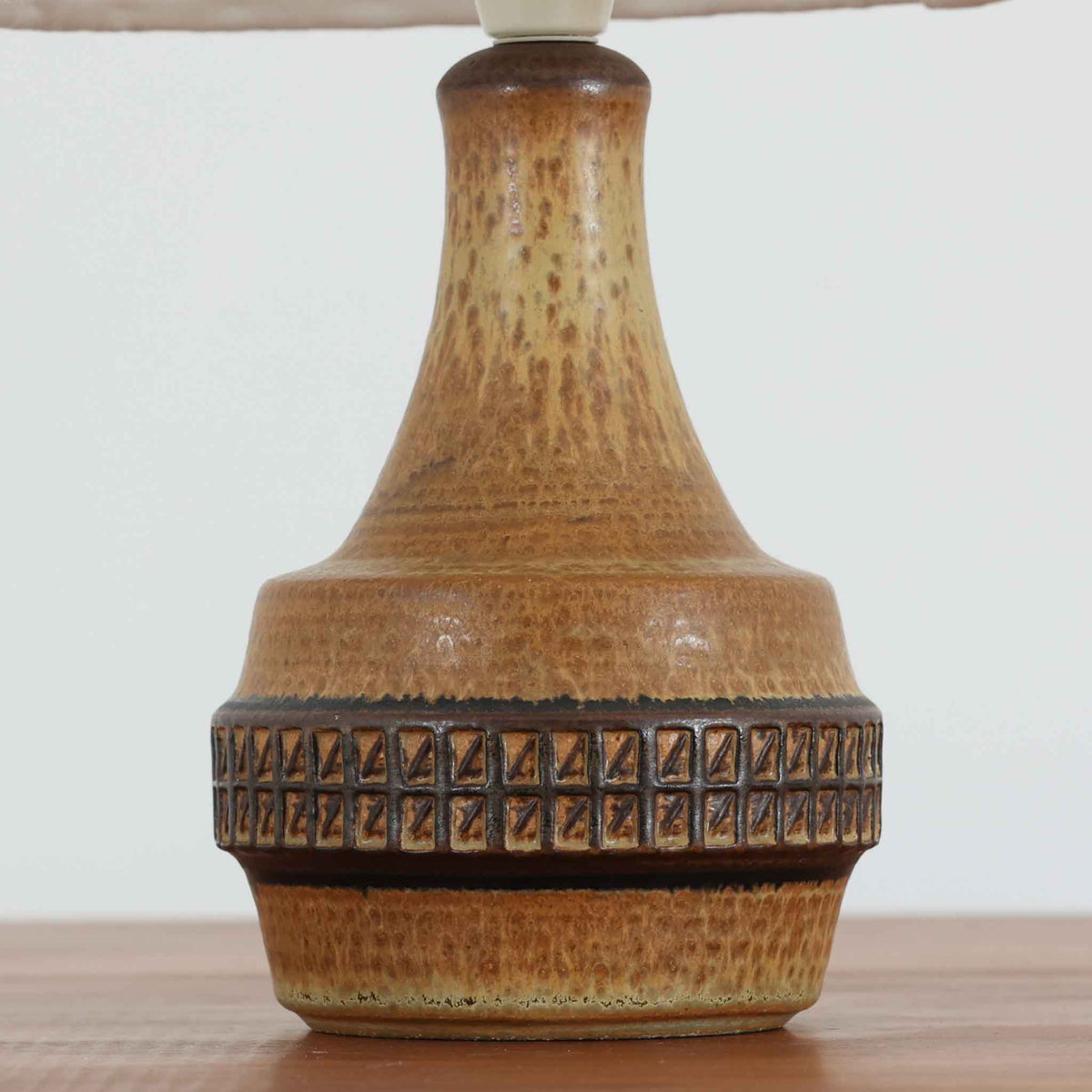 デンマークの陶磁器メーカーSoholm Keramik（スーホルム）による、Joseph Simon（ヨゼフ・シモン）デザインのビンテージテーブルランプ。温かみのある琥珀色の釉薬が施された陶磁器製ベースは、細長く伸びた首元から裾に向かって広がる優美なフォルムが特徴です。下部には幾何学模様のレリーフが帯状に施され、釉薬の濃淡と相まって立体感のある豊かな表情を生み出しています。素朴な風合いの陶器と、繊細なプリーツを施した白いリネンシェードの組み合わせも魅力的。点灯すると布越しに柔らかな光が広がり、落ち着いた雰囲気を演出します。北欧のクラフトマンシップが感じられる味わい深い一台で、リビングや寝室のアクセント照明としておすすめです。