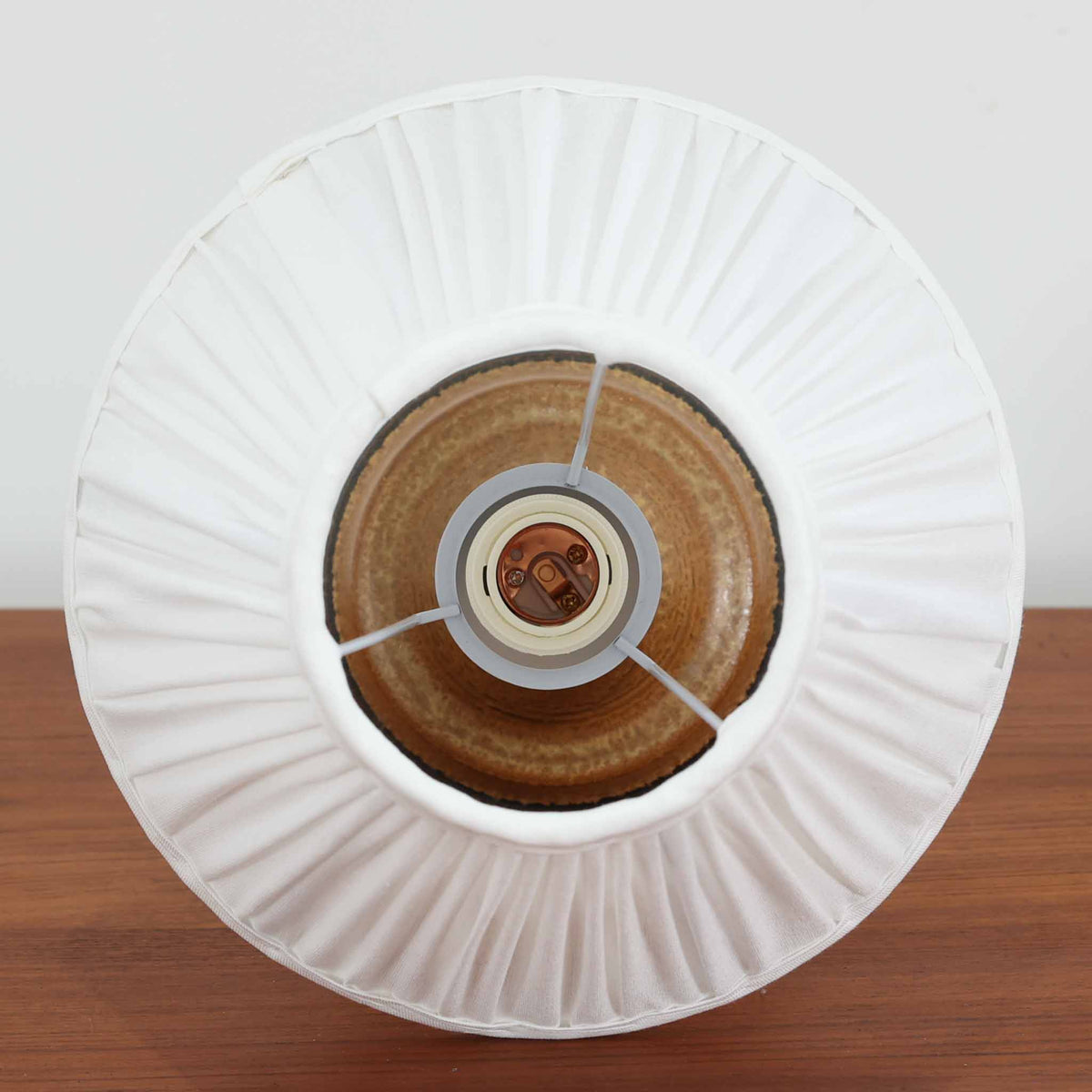 デンマークの陶磁器メーカーSoholm Keramik（スーホルム）による、Joseph Simon（ヨゼフ・シモン）デザインのビンテージテーブルランプ。温かみのある琥珀色の釉薬が施された陶磁器製ベースは、細長く伸びた首元から裾に向かって広がる優美なフォルムが特徴です。下部には幾何学模様のレリーフが帯状に施され、釉薬の濃淡と相まって立体感のある豊かな表情を生み出しています。素朴な風合いの陶器と、繊細なプリーツを施した白いリネンシェードの組み合わせも魅力的。点灯すると布越しに柔らかな光が広がり、落ち着いた雰囲気を演出します。北欧のクラフトマンシップが感じられる味わい深い一台で、リビングや寝室のアクセント照明としておすすめです。