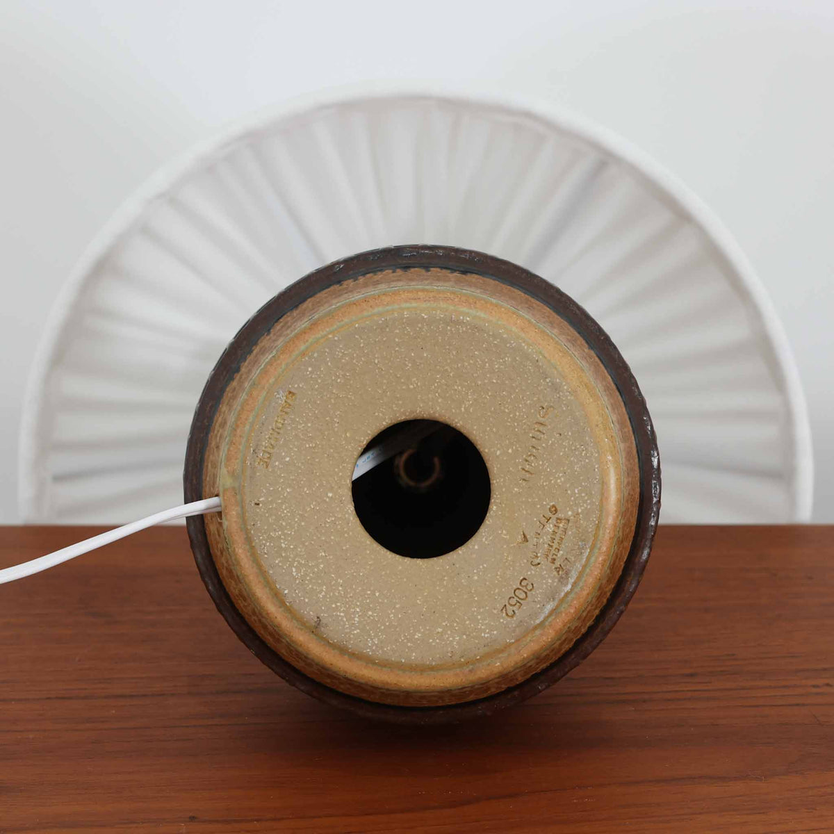 デンマークの陶磁器メーカーSoholm Keramik（スーホルム）による、Joseph Simon（ヨゼフ・シモン）デザインのビンテージテーブルランプ。温かみのある琥珀色の釉薬が施された陶磁器製ベースは、細長く伸びた首元から裾に向かって広がる優美なフォルムが特徴です。下部には幾何学模様のレリーフが帯状に施され、釉薬の濃淡と相まって立体感のある豊かな表情を生み出しています。素朴な風合いの陶器と、繊細なプリーツを施した白いリネンシェードの組み合わせも魅力的。点灯すると布越しに柔らかな光が広がり、落ち着いた雰囲気を演出します。北欧のクラフトマンシップが感じられる味わい深い一台で、リビングや寝室のアクセント照明としておすすめです。