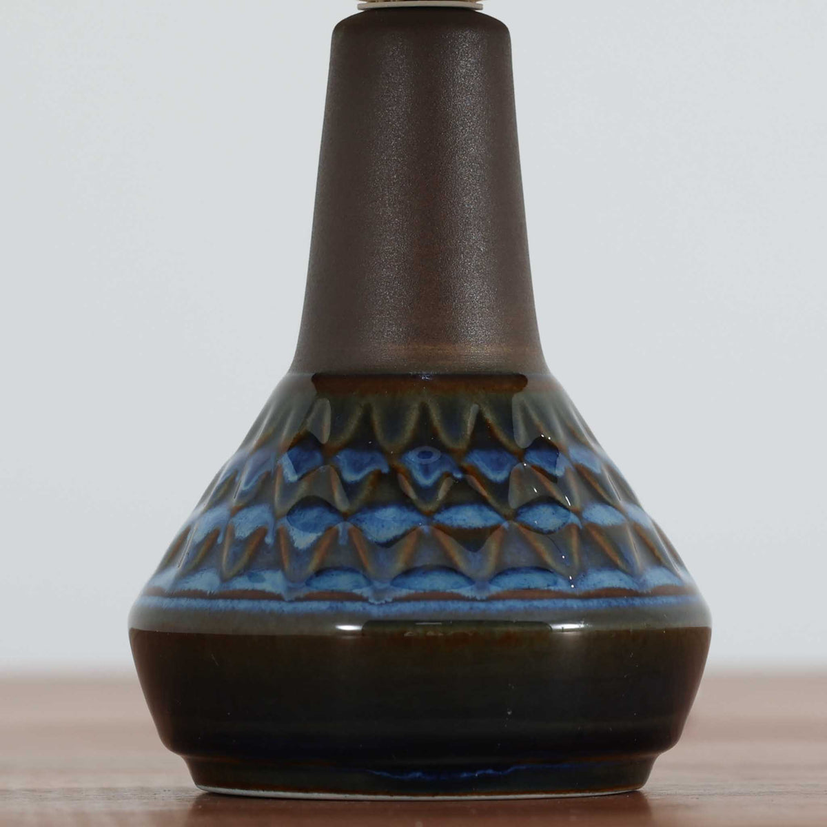 デンマークの陶磁器メーカーSoholm Keramik（スーホルム）による、Einar Johansen（アイナー・ヨハンセン）デザインのビンテージテーブルランプ。すっきりと伸びた首元から裾に向かって広がる陶磁器製ベースは、落ち着いたブラウンと鮮やかなブルーの釉薬が織りなす美しい色彩が特徴です。胴部には連続する幾何学模様のレリーフが施され、釉薬の濃淡と立体的な陰影が豊かな表情を生み出しています。素朴な陶器の質感と、繊細なプリーツを施した白いリネンシェードの組み合わせも魅力的。点灯すると布越しに柔らかな光が広がり、空間を温かく包み込みます。北欧らしい造形美とクラフトマンシップが感じられる一台で、リビングや寝室のアクセント照明としておすすめです。
