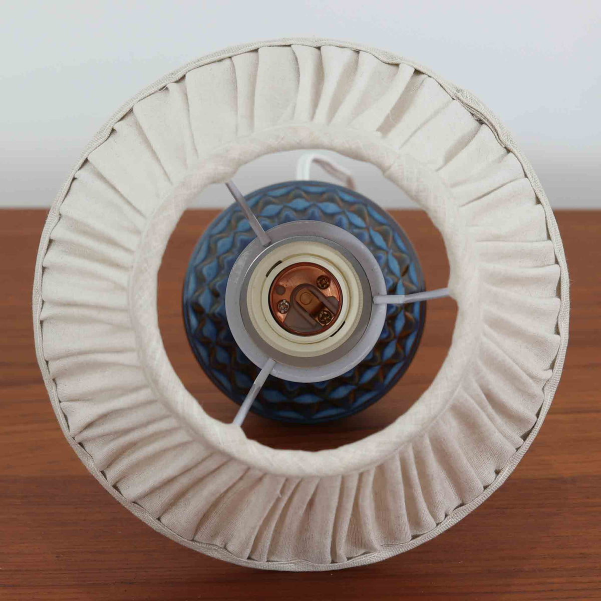 デンマークの陶磁器メーカーSoholm Keramik（スーホルム）による、Einar Johansen（アイナー・ヨハンセン）デザインのビンテージテーブルランプ。すっきりと伸びた首元から裾に向かって広がる陶磁器製ベースは、落ち着いたブラウンと鮮やかなブルーの釉薬が織りなす美しい色彩が特徴です。胴部には連続する幾何学模様のレリーフが施され、釉薬の濃淡と立体的な陰影が豊かな表情を生み出しています。素朴な陶器の質感と、繊細なプリーツを施した白いリネンシェードの組み合わせも魅力的。点灯すると布越しに柔らかな光が広がり、空間を温かく包み込みます。北欧らしい造形美とクラフトマンシップが感じられる一台で、リビングや寝室のアクセント照明としておすすめです。
