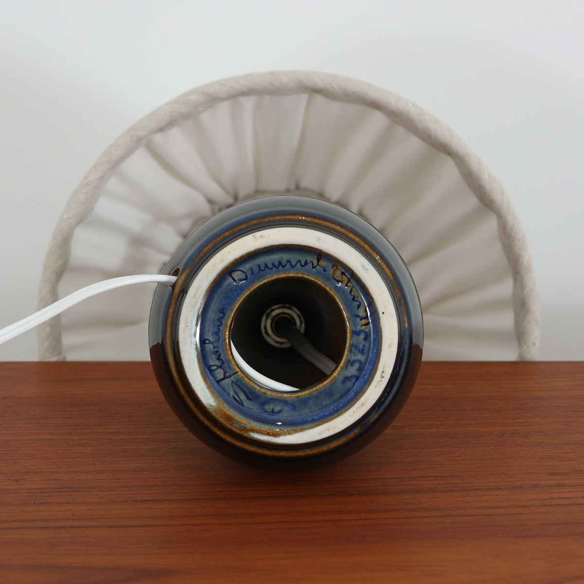 デンマークの陶磁器メーカーSoholm Keramik（スーホルム）による、Einar Johansen（アイナー・ヨハンセン）デザインのビンテージテーブルランプ。すっきりと伸びた首元から裾に向かって広がる陶磁器製ベースは、落ち着いたブラウンと鮮やかなブルーの釉薬が織りなす美しい色彩が特徴です。胴部には連続する幾何学模様のレリーフが施され、釉薬の濃淡と立体的な陰影が豊かな表情を生み出しています。素朴な陶器の質感と、繊細なプリーツを施した白いリネンシェードの組み合わせも魅力的。点灯すると布越しに柔らかな光が広がり、空間を温かく包み込みます。北欧らしい造形美とクラフトマンシップが感じられる一台で、リビングや寝室のアクセント照明としておすすめです。