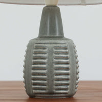 デンマークの陶磁器メーカーSoholm Keramik（スーホルム）による、Einar Johansen（アイナー・ヨハンセン）デザインのビンテージテーブルランプ。落ち着いたグレイッシュグリーンの釉薬が施された陶磁器製ベースは、すっきりと伸びた首元と丸みを帯びた胴部のバランスが美しい、端正なフォルムが特徴です。胴部には横方向に連なる溝状のレリーフが規則的に施され、滑らかな面とのコントラストが立体感のある表情を生み出しています。釉薬の繊細な濃淡や陰影には、ビンテージ陶器ならではの味わいが感じられます。シンプルな白いリネンシェードを組み合わせることで、重厚感のあるベースに軽やかさを添え、点灯時には柔らかな光が空間に広がります。リビングや寝室のアクセント照明としておすすめの一台です。