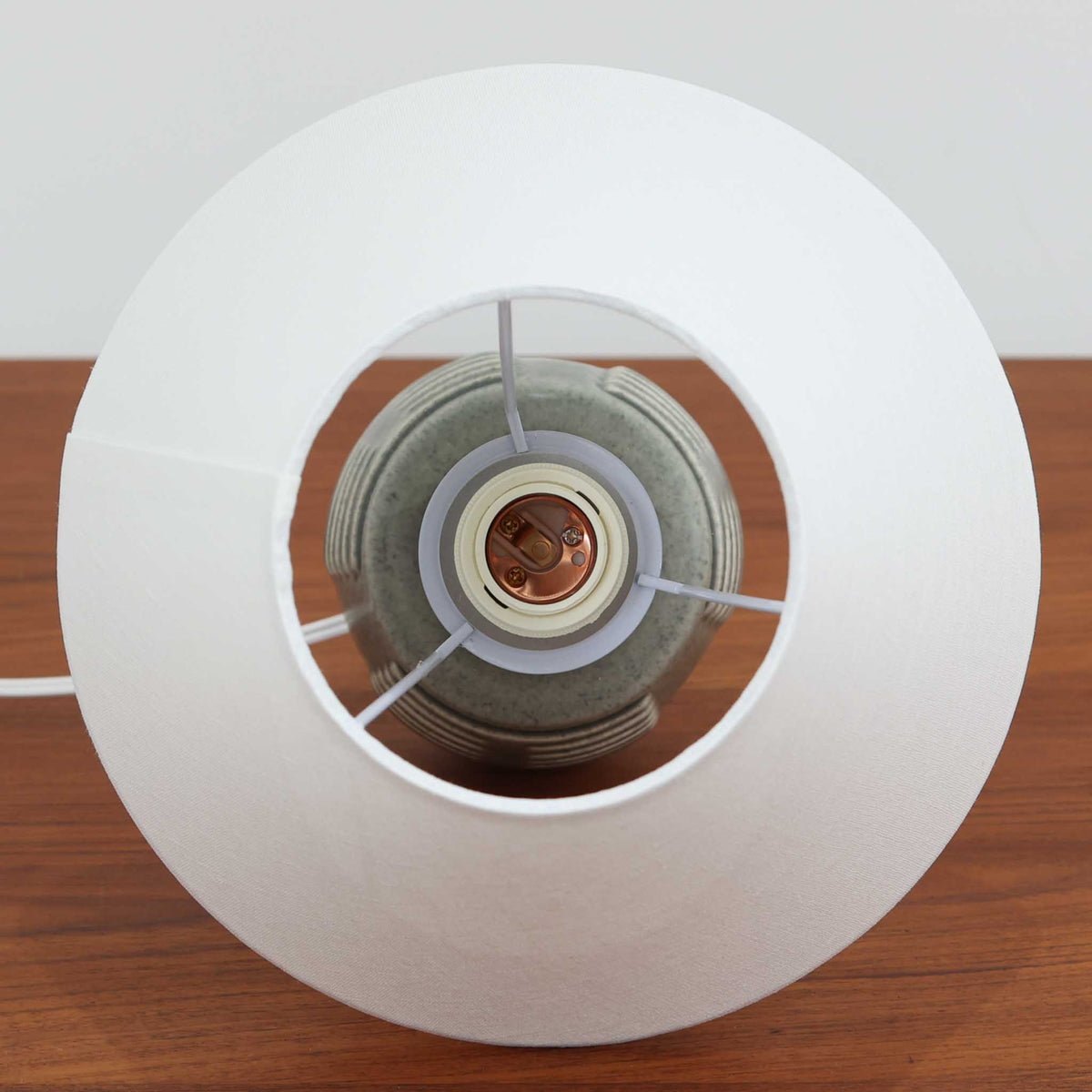 デンマークの陶磁器メーカーSoholm Keramik（スーホルム）による、Einar Johansen（アイナー・ヨハンセン）デザインのビンテージテーブルランプ。落ち着いたグレイッシュグリーンの釉薬が施された陶磁器製ベースは、すっきりと伸びた首元と丸みを帯びた胴部のバランスが美しい、端正なフォルムが特徴です。胴部には横方向に連なる溝状のレリーフが規則的に施され、滑らかな面とのコントラストが立体感のある表情を生み出しています。釉薬の繊細な濃淡や陰影には、ビンテージ陶器ならではの味わいが感じられます。シンプルな白いリネンシェードを組み合わせることで、重厚感のあるベースに軽やかさを添え、点灯時には柔らかな光が空間に広がります。リビングや寝室のアクセント照明としておすすめの一台です。