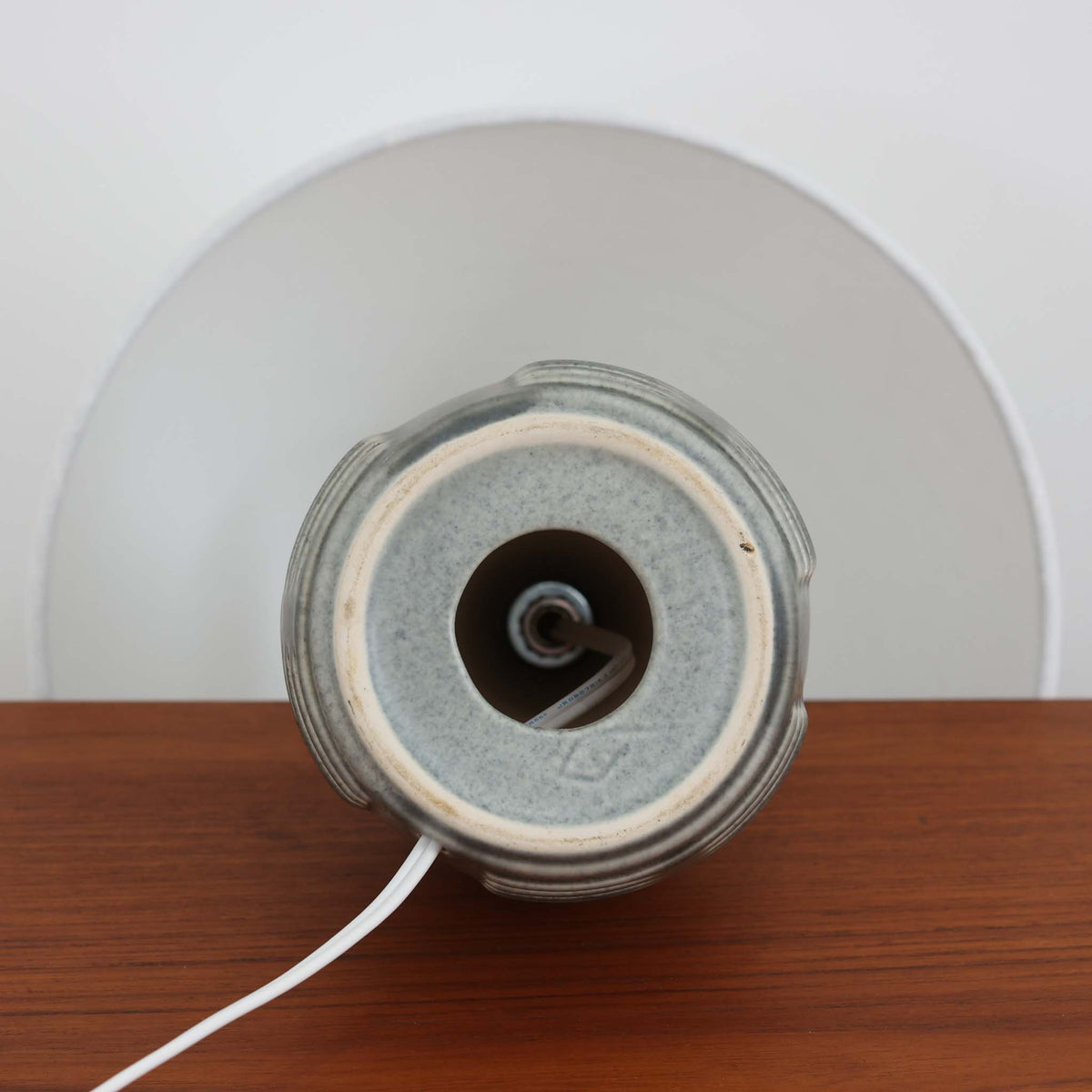 デンマークの陶磁器メーカーSoholm Keramik（スーホルム）による、Einar Johansen（アイナー・ヨハンセン）デザインのビンテージテーブルランプ。落ち着いたグレイッシュグリーンの釉薬が施された陶磁器製ベースは、すっきりと伸びた首元と丸みを帯びた胴部のバランスが美しい、端正なフォルムが特徴です。胴部には横方向に連なる溝状のレリーフが規則的に施され、滑らかな面とのコントラストが立体感のある表情を生み出しています。釉薬の繊細な濃淡や陰影には、ビンテージ陶器ならではの味わいが感じられます。シンプルな白いリネンシェードを組み合わせることで、重厚感のあるベースに軽やかさを添え、点灯時には柔らかな光が空間に広がります。リビングや寝室のアクセント照明としておすすめの一台です。
