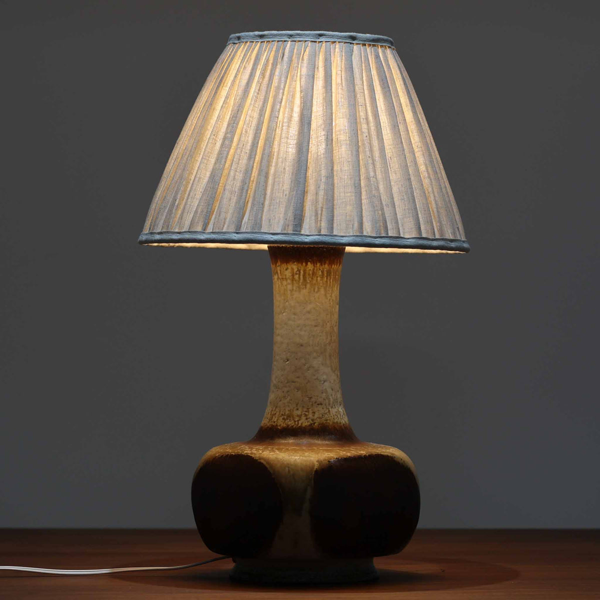 デンマークの陶磁器メーカーAxella Keramik（アクセラ）による、Jette Helleroe（イェッテ・ヘレロー）デザインのビンテージテーブルランプ。すらりと伸びた細身の首元と、ふっくらと丸みを帯びたベースが印象的な、美しいシルエットの一台です。陶磁器製の本体には、アイボリーを基調とした柔らかな色合いに、深みのあるブラウンの釉薬が大胆に施され、流れるような色の境界や自然な濃淡が豊かな表情を生み出しています。釉薬の重なりによって生まれる独特の風合いには、手仕事ならではの温もりと芸術性が感じられます。プリーツを施したリネンシェードは柔らかな光を広げ、存在感のあるベースとの美しい調和を演出します。リビングのサイドボードや寝室など、空間を印象的に彩るアクセント照明としておすすめです。