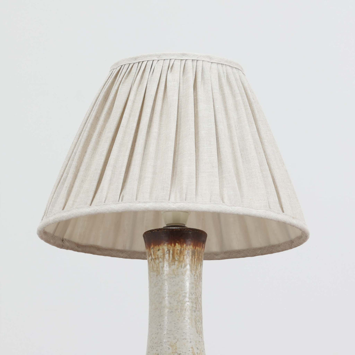 デンマークの陶磁器メーカーAxella Keramik（アクセラ）による、Jette Helleroe（イェッテ・ヘレロー）デザインのビンテージテーブルランプ。すらりと伸びた細身の首元と、ふっくらと丸みを帯びたベースが印象的な、美しいシルエットの一台です。陶磁器製の本体には、アイボリーを基調とした柔らかな色合いに、深みのあるブラウンの釉薬が大胆に施され、流れるような色の境界や自然な濃淡が豊かな表情を生み出しています。釉薬の重なりによって生まれる独特の風合いには、手仕事ならではの温もりと芸術性が感じられます。プリーツを施したリネンシェードは柔らかな光を広げ、存在感のあるベースとの美しい調和を演出します。リビングのサイドボードや寝室など、空間を印象的に彩るアクセント照明としておすすめです。