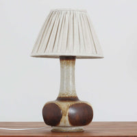 デンマークの陶磁器メーカーAxella Keramik（アクセラ）による、Jette Helleroe（イェッテ・ヘレロー）デザインのビンテージテーブルランプ。すらりと伸びた細身の首元と、ふっくらと丸みを帯びたベースが印象的な、美しいシルエットの一台です。陶磁器製の本体には、アイボリーを基調とした柔らかな色合いに、深みのあるブラウンの釉薬が大胆に施され、流れるような色の境界や自然な濃淡が豊かな表情を生み出しています。釉薬の重なりによって生まれる独特の風合いには、手仕事ならではの温もりと芸術性が感じられます。プリーツを施したリネンシェードは柔らかな光を広げ、存在感のあるベースとの美しい調和を演出します。リビングのサイドボードや寝室など、空間を印象的に彩るアクセント照明としておすすめです。