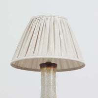 デンマークの陶磁器メーカーAxella Keramik（アクセラ）による、Jette Helleroe（イェッテ・ヘレロー）デザインのビンテージテーブルランプ。すらりと伸びた細身の首元と、ふっくらと丸みを帯びたベースが印象的な、美しいシルエットの一台です。陶磁器製の本体には、アイボリーを基調とした柔らかな色合いに、深みのあるブラウンの釉薬が大胆に施され、流れるような色の境界や自然な濃淡が豊かな表情を生み出しています。釉薬の重なりによって生まれる独特の風合いには、手仕事ならではの温もりと芸術性が感じられます。プリーツを施したリネンシェードは柔らかな光を広げ、存在感のあるベースとの美しい調和を演出します。リビングのサイドボードや寝室など、空間を印象的に彩るアクセント照明としておすすめです。