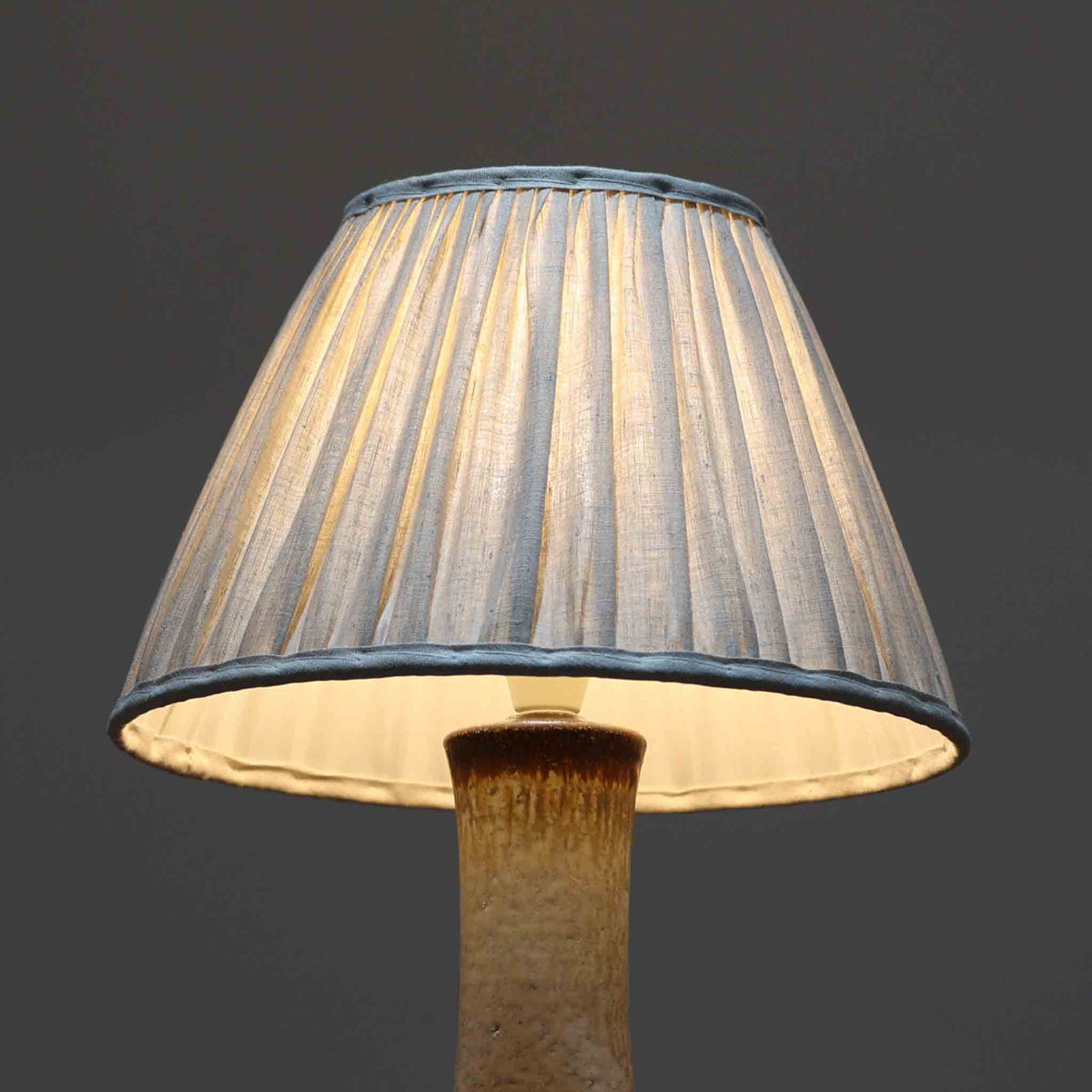 デンマークの陶磁器メーカーAxella Keramik（アクセラ）による、Jette Helleroe（イェッテ・ヘレロー）デザインのビンテージテーブルランプ。すらりと伸びた細身の首元と、ふっくらと丸みを帯びたベースが印象的な、美しいシルエットの一台です。陶磁器製の本体には、アイボリーを基調とした柔らかな色合いに、深みのあるブラウンの釉薬が大胆に施され、流れるような色の境界や自然な濃淡が豊かな表情を生み出しています。釉薬の重なりによって生まれる独特の風合いには、手仕事ならではの温もりと芸術性が感じられます。プリーツを施したリネンシェードは柔らかな光を広げ、存在感のあるベースとの美しい調和を演出します。リビングのサイドボードや寝室など、空間を印象的に彩るアクセント照明としておすすめです。