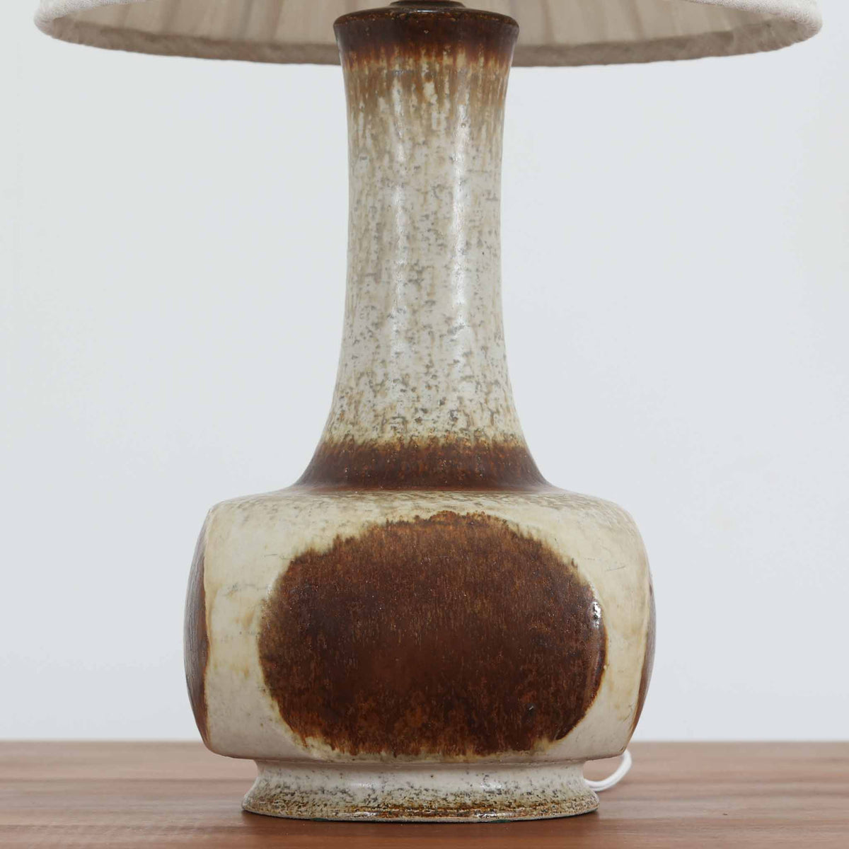 デンマークの陶磁器メーカーAxella Keramik（アクセラ）による、Jette Helleroe（イェッテ・ヘレロー）デザインのビンテージテーブルランプ。すらりと伸びた細身の首元と、ふっくらと丸みを帯びたベースが印象的な、美しいシルエットの一台です。陶磁器製の本体には、アイボリーを基調とした柔らかな色合いに、深みのあるブラウンの釉薬が大胆に施され、流れるような色の境界や自然な濃淡が豊かな表情を生み出しています。釉薬の重なりによって生まれる独特の風合いには、手仕事ならではの温もりと芸術性が感じられます。プリーツを施したリネンシェードは柔らかな光を広げ、存在感のあるベースとの美しい調和を演出します。リビングのサイドボードや寝室など、空間を印象的に彩るアクセント照明としておすすめです。