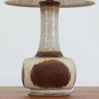デンマークの陶磁器メーカーAxella Keramik（アクセラ）による、Jette Helleroe（イェッテ・ヘレロー）デザインのビンテージテーブルランプ。すらりと伸びた細身の首元と、ふっくらと丸みを帯びたベースが印象的な、美しいシルエットの一台です。陶磁器製の本体には、アイボリーを基調とした柔らかな色合いに、深みのあるブラウンの釉薬が大胆に施され、流れるような色の境界や自然な濃淡が豊かな表情を生み出しています。釉薬の重なりによって生まれる独特の風合いには、手仕事ならではの温もりと芸術性が感じられます。プリーツを施したリネンシェードは柔らかな光を広げ、存在感のあるベースとの美しい調和を演出します。リビングのサイドボードや寝室など、空間を印象的に彩るアクセント照明としておすすめです。