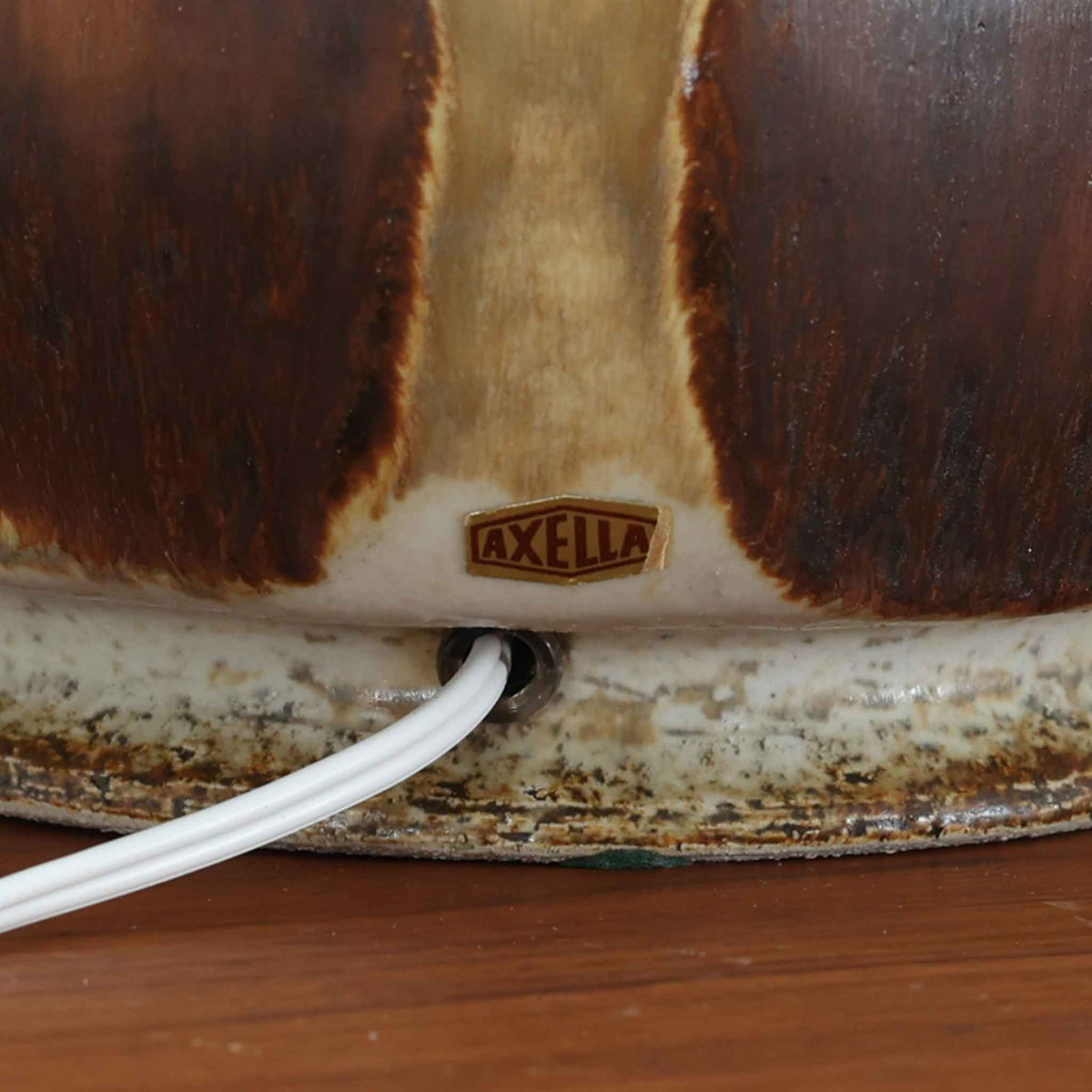 デンマークの陶磁器メーカーAxella Keramik（アクセラ）による、Jette Helleroe（イェッテ・ヘレロー）デザインのビンテージテーブルランプ。すらりと伸びた細身の首元と、ふっくらと丸みを帯びたベースが印象的な、美しいシルエットの一台です。陶磁器製の本体には、アイボリーを基調とした柔らかな色合いに、深みのあるブラウンの釉薬が大胆に施され、流れるような色の境界や自然な濃淡が豊かな表情を生み出しています。釉薬の重なりによって生まれる独特の風合いには、手仕事ならではの温もりと芸術性が感じられます。プリーツを施したリネンシェードは柔らかな光を広げ、存在感のあるベースとの美しい調和を演出します。リビングのサイドボードや寝室など、空間を印象的に彩るアクセント照明としておすすめです。