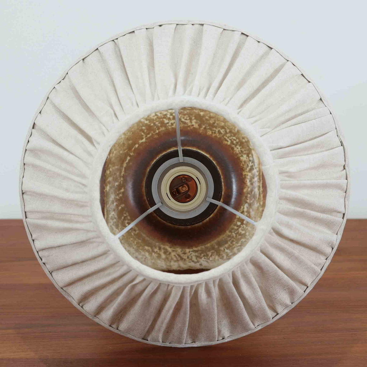 デンマークの陶磁器メーカーAxella Keramik（アクセラ）による、Jette Helleroe（イェッテ・ヘレロー）デザインのビンテージテーブルランプ。すらりと伸びた細身の首元と、ふっくらと丸みを帯びたベースが印象的な、美しいシルエットの一台です。陶磁器製の本体には、アイボリーを基調とした柔らかな色合いに、深みのあるブラウンの釉薬が大胆に施され、流れるような色の境界や自然な濃淡が豊かな表情を生み出しています。釉薬の重なりによって生まれる独特の風合いには、手仕事ならではの温もりと芸術性が感じられます。プリーツを施したリネンシェードは柔らかな光を広げ、存在感のあるベースとの美しい調和を演出します。リビングのサイドボードや寝室など、空間を印象的に彩るアクセント照明としておすすめです。