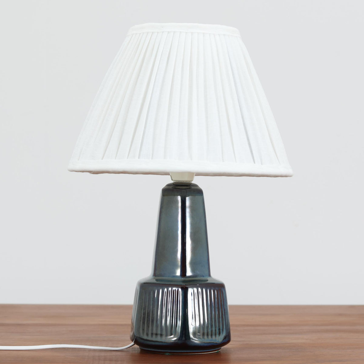 デンマークの陶磁器メーカーSoholm Keramik（スーホルム）による、Einar Johansen（アイナー・ヨハンセン）デザインのビンテージテーブルランプ。すらりと伸びた首元と、裾に向かって緩やかに広がるベースが美しい、端正で洗練されたシルエットが特徴です。深みのあるブルーグレーの釉薬は、光の当たり方によって繊細な色の変化を見せ、艶やかな質感が陶磁器ならではの魅力を引き立てています。ベース下部には縦方向のレリーフ模様が規則的に施され、立体的な陰影と心地よいリズムを生み出しています。プリーツを施したリネンシェードからは柔らかな光が広がり、落ち着いた雰囲気を演出します。リビングのサイドボードや寝室など、空間に上品なアクセントを添える照明としておすすめです。