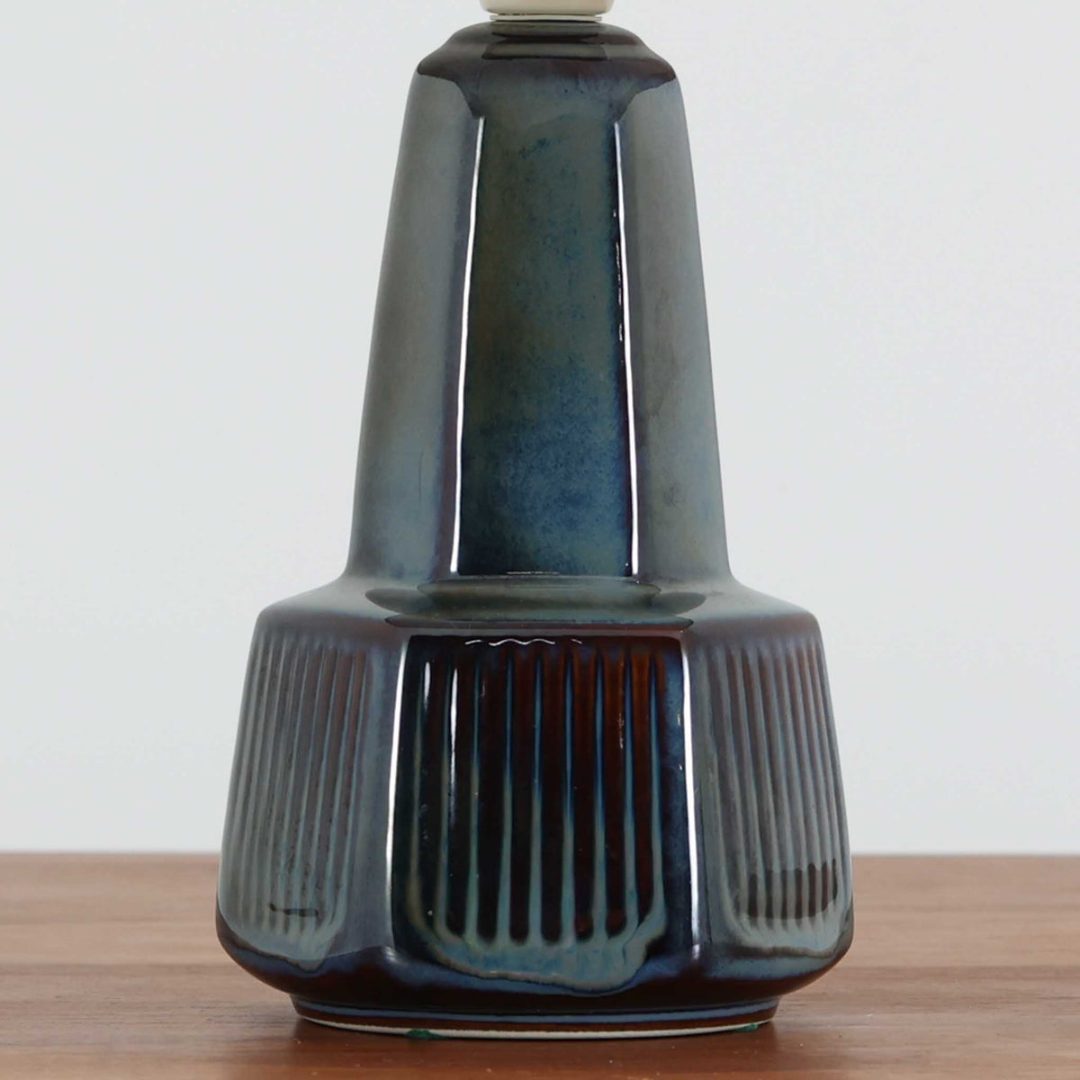 デンマークの陶磁器メーカーSoholm Keramik（スーホルム）による、Einar Johansen（アイナー・ヨハンセン）デザインのビンテージテーブルランプ。すらりと伸びた首元と、裾に向かって緩やかに広がるベースが美しい、端正で洗練されたシルエットが特徴です。深みのあるブルーグレーの釉薬は、光の当たり方によって繊細な色の変化を見せ、艶やかな質感が陶磁器ならではの魅力を引き立てています。ベース下部には縦方向のレリーフ模様が規則的に施され、立体的な陰影と心地よいリズムを生み出しています。プリーツを施したリネンシェードからは柔らかな光が広がり、落ち着いた雰囲気を演出します。リビングのサイドボードや寝室など、空間に上品なアクセントを添える照明としておすすめです。