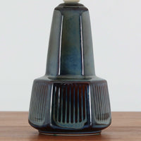 デンマークの陶磁器メーカーSoholm Keramik（スーホルム）による、Einar Johansen（アイナー・ヨハンセン）デザインのビンテージテーブルランプ。すらりと伸びた首元と、裾に向かって緩やかに広がるベースが美しい、端正で洗練されたシルエットが特徴です。深みのあるブルーグレーの釉薬は、光の当たり方によって繊細な色の変化を見せ、艶やかな質感が陶磁器ならではの魅力を引き立てています。ベース下部には縦方向のレリーフ模様が規則的に施され、立体的な陰影と心地よいリズムを生み出しています。プリーツを施したリネンシェードからは柔らかな光が広がり、落ち着いた雰囲気を演出します。リビングのサイドボードや寝室など、空間に上品なアクセントを添える照明としておすすめです。
