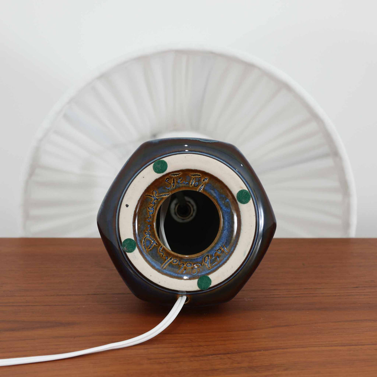 デンマークの陶磁器メーカーSoholm Keramik（スーホルム）による、Einar Johansen（アイナー・ヨハンセン）デザインのビンテージテーブルランプ。すらりと伸びた首元と、裾に向かって緩やかに広がるベースが美しい、端正で洗練されたシルエットが特徴です。深みのあるブルーグレーの釉薬は、光の当たり方によって繊細な色の変化を見せ、艶やかな質感が陶磁器ならではの魅力を引き立てています。ベース下部には縦方向のレリーフ模様が規則的に施され、立体的な陰影と心地よいリズムを生み出しています。プリーツを施したリネンシェードからは柔らかな光が広がり、落ち着いた雰囲気を演出します。リビングのサイドボードや寝室など、空間に上品なアクセントを添える照明としておすすめです。