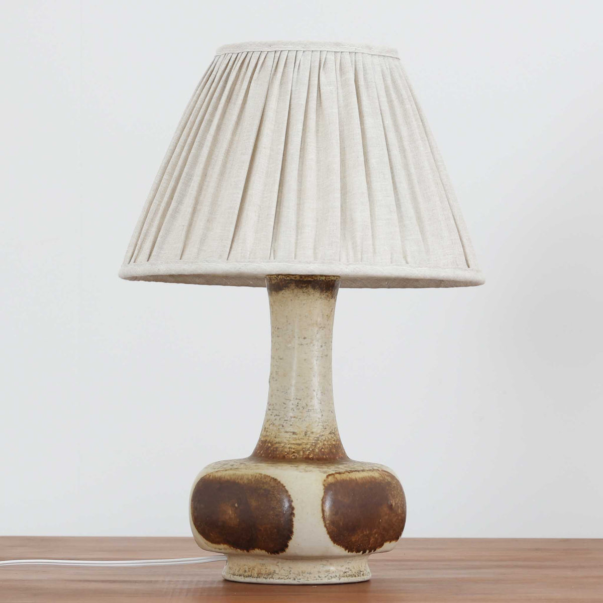 デンマークの陶磁器メーカーAxella Keramik（アクセラ）による、Jette Helleroe（イェッテ・ヘレロー）デザインのビンテージテーブルランプ。丸みを帯びたふっくらとしたベースから、細く伸びる首元へとつながる彫刻的なフォルムが印象的な一品です。温かみのあるアイボリーを基調に、深いブラウンの釉薬を大胆に配した独特の模様が特徴で、釉薬の自然な濃淡や境界のにじみが、手仕事ならではの豊かな表情を生み出しています。素朴さと芸術性を兼ね備えた佇まいは、北欧ビンテージならではの魅力を感じさせます。プリーツを施したリネンシェードは柔らかな光を広げ、落ち着いた温もりのある空間を演出します。リビングのサイドボードや寝室のチェストなどに置けば、消灯時にもオブジェのような存在感を楽しめます。