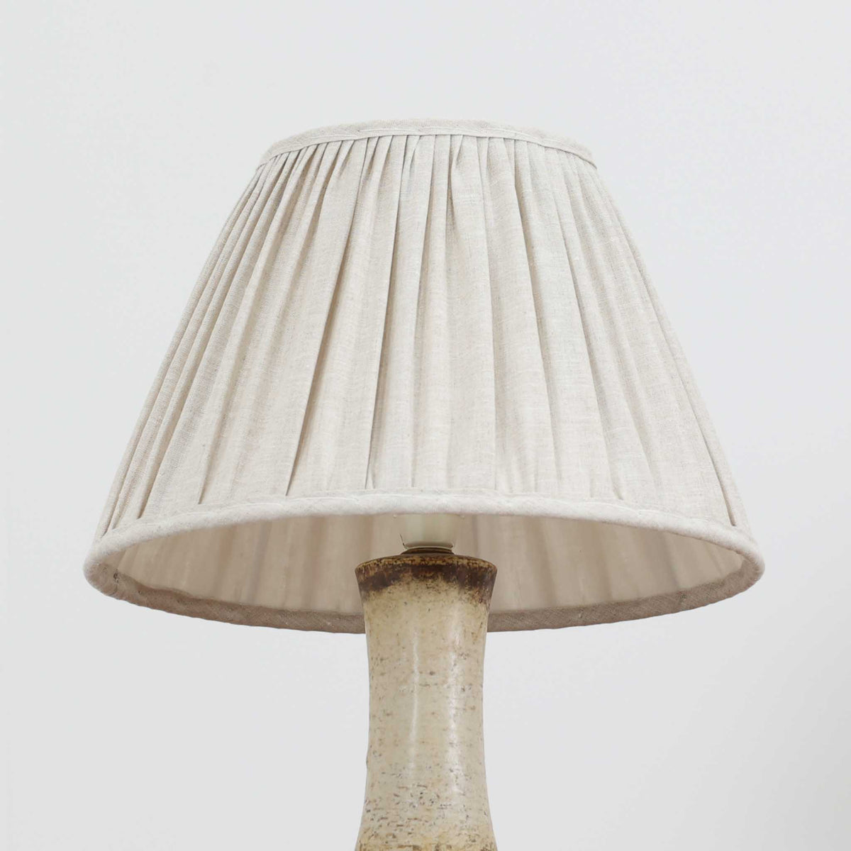 デンマークの陶磁器メーカーAxella Keramik（アクセラ）による、Jette Helleroe（イェッテ・ヘレロー）デザインのビンテージテーブルランプ。丸みを帯びたふっくらとしたベースから、細く伸びる首元へとつながる彫刻的なフォルムが印象的な一品です。温かみのあるアイボリーを基調に、深いブラウンの釉薬を大胆に配した独特の模様が特徴で、釉薬の自然な濃淡や境界のにじみが、手仕事ならではの豊かな表情を生み出しています。素朴さと芸術性を兼ね備えた佇まいは、北欧ビンテージならではの魅力を感じさせます。プリーツを施したリネンシェードは柔らかな光を広げ、落ち着いた温もりのある空間を演出します。リビングのサイドボードや寝室のチェストなどに置けば、消灯時にもオブジェのような存在感を楽しめます。