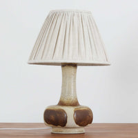 デンマークの陶磁器メーカーAxella Keramik（アクセラ）による、Jette Helleroe（イェッテ・ヘレロー）デザインのビンテージテーブルランプ。丸みを帯びたふっくらとしたベースから、細く伸びる首元へとつながる彫刻的なフォルムが印象的な一品です。温かみのあるアイボリーを基調に、深いブラウンの釉薬を大胆に配した独特の模様が特徴で、釉薬の自然な濃淡や境界のにじみが、手仕事ならではの豊かな表情を生み出しています。素朴さと芸術性を兼ね備えた佇まいは、北欧ビンテージならではの魅力を感じさせます。プリーツを施したリネンシェードは柔らかな光を広げ、落ち着いた温もりのある空間を演出します。リビングのサイドボードや寝室のチェストなどに置けば、消灯時にもオブジェのような存在感を楽しめます。