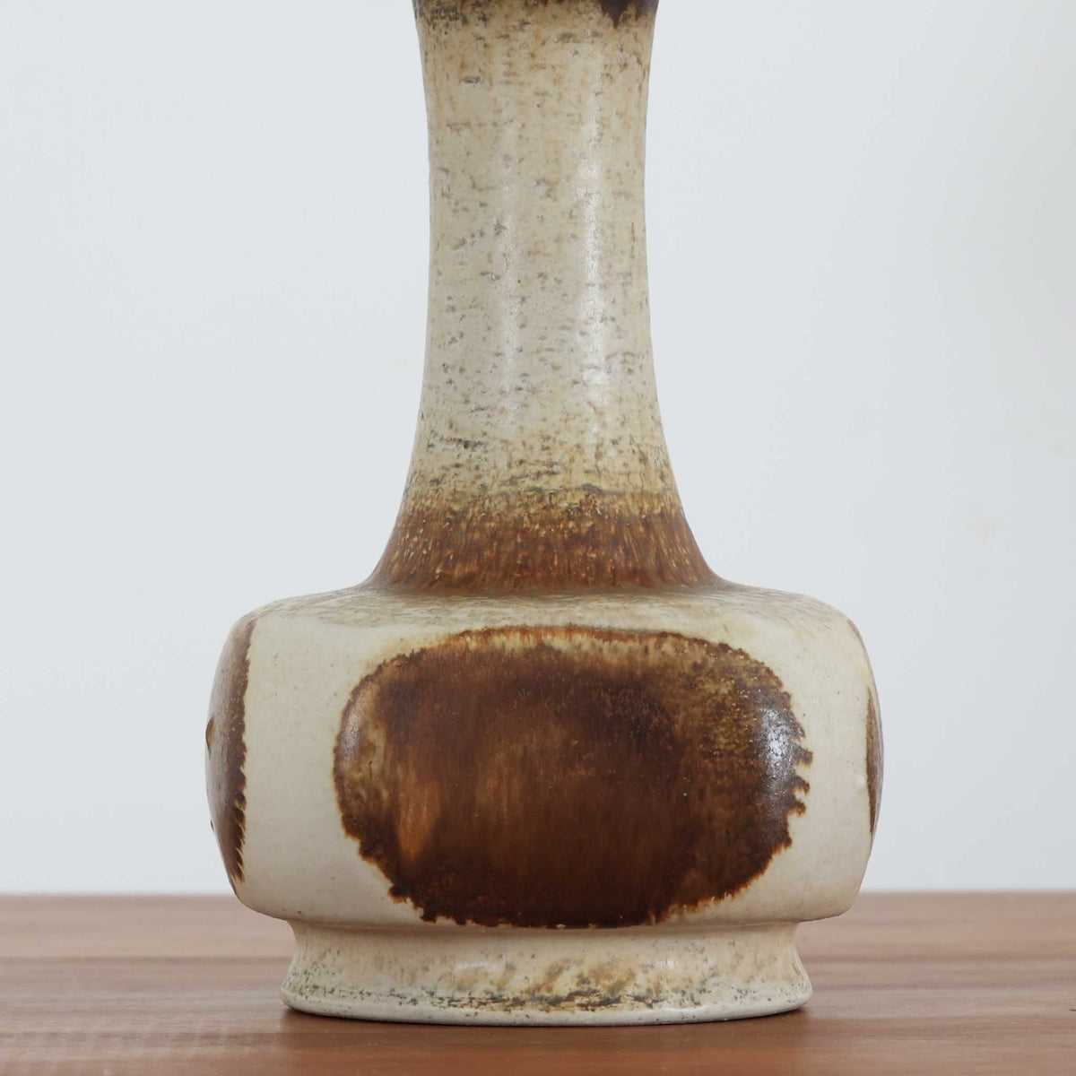 デンマークの陶磁器メーカーAxella Keramik（アクセラ）による、Jette Helleroe（イェッテ・ヘレロー）デザインのビンテージテーブルランプ。丸みを帯びたふっくらとしたベースから、細く伸びる首元へとつながる彫刻的なフォルムが印象的な一品です。温かみのあるアイボリーを基調に、深いブラウンの釉薬を大胆に配した独特の模様が特徴で、釉薬の自然な濃淡や境界のにじみが、手仕事ならではの豊かな表情を生み出しています。素朴さと芸術性を兼ね備えた佇まいは、北欧ビンテージならではの魅力を感じさせます。プリーツを施したリネンシェードは柔らかな光を広げ、落ち着いた温もりのある空間を演出します。リビングのサイドボードや寝室のチェストなどに置けば、消灯時にもオブジェのような存在感を楽しめます。