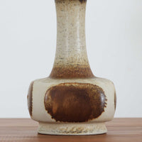 デンマークの陶磁器メーカーAxella Keramik（アクセラ）による、Jette Helleroe（イェッテ・ヘレロー）デザインのビンテージテーブルランプ。丸みを帯びたふっくらとしたベースから、細く伸びる首元へとつながる彫刻的なフォルムが印象的な一品です。温かみのあるアイボリーを基調に、深いブラウンの釉薬を大胆に配した独特の模様が特徴で、釉薬の自然な濃淡や境界のにじみが、手仕事ならではの豊かな表情を生み出しています。素朴さと芸術性を兼ね備えた佇まいは、北欧ビンテージならではの魅力を感じさせます。プリーツを施したリネンシェードは柔らかな光を広げ、落ち着いた温もりのある空間を演出します。リビングのサイドボードや寝室のチェストなどに置けば、消灯時にもオブジェのような存在感を楽しめます。