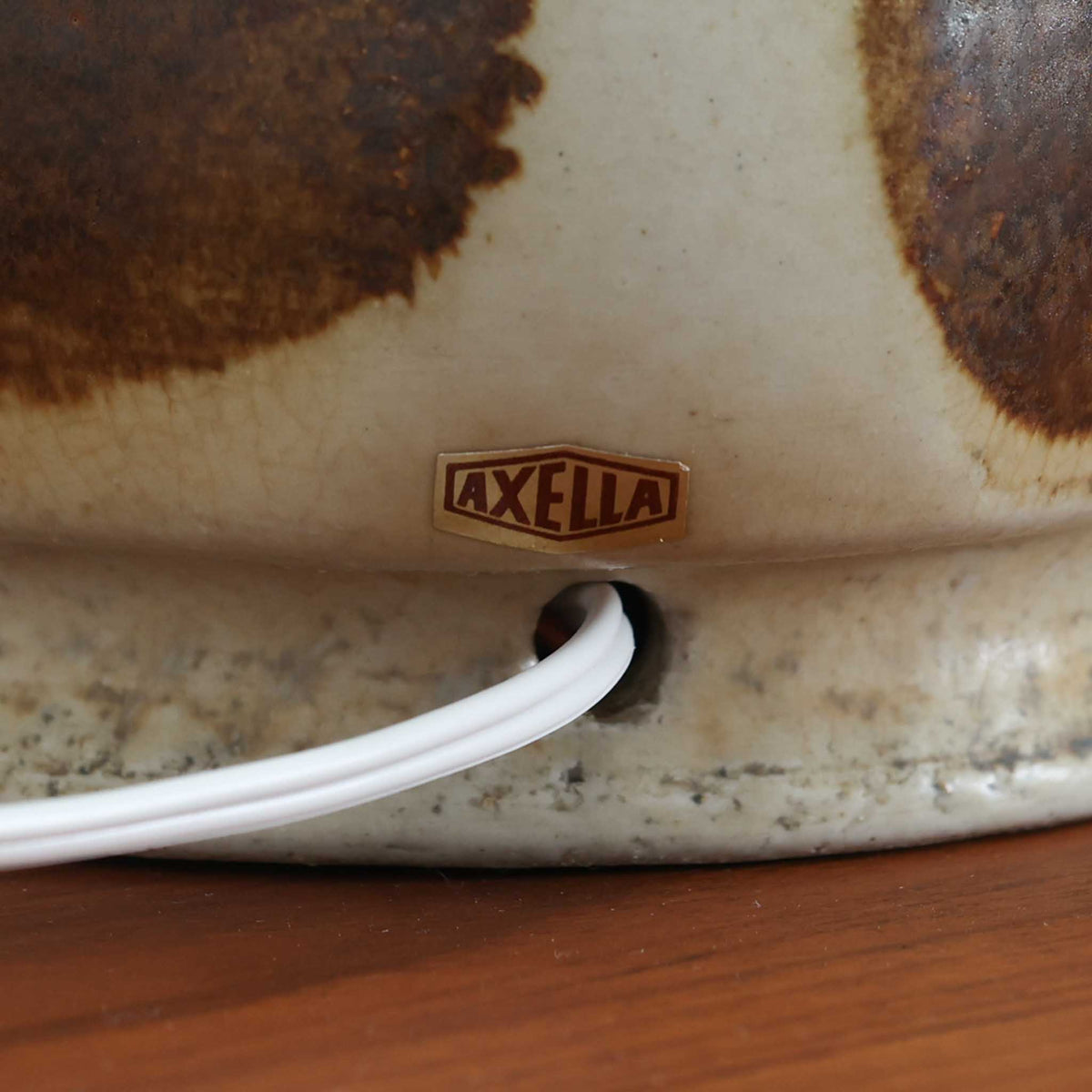 デンマークの陶磁器メーカーAxella Keramik（アクセラ）による、Jette Helleroe（イェッテ・ヘレロー）デザインのビンテージテーブルランプ。丸みを帯びたふっくらとしたベースから、細く伸びる首元へとつながる彫刻的なフォルムが印象的な一品です。温かみのあるアイボリーを基調に、深いブラウンの釉薬を大胆に配した独特の模様が特徴で、釉薬の自然な濃淡や境界のにじみが、手仕事ならではの豊かな表情を生み出しています。素朴さと芸術性を兼ね備えた佇まいは、北欧ビンテージならではの魅力を感じさせます。プリーツを施したリネンシェードは柔らかな光を広げ、落ち着いた温もりのある空間を演出します。リビングのサイドボードや寝室のチェストなどに置けば、消灯時にもオブジェのような存在感を楽しめます。