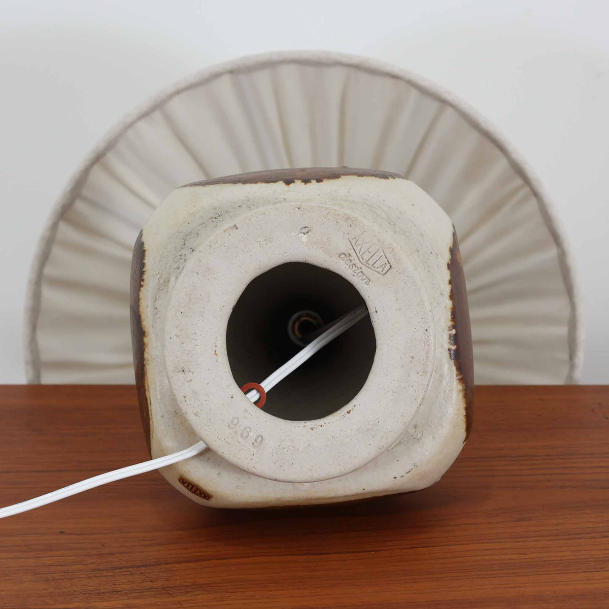 デンマークの陶磁器メーカーAxella Keramik（アクセラ）による、Jette Helleroe（イェッテ・ヘレロー）デザインのビンテージテーブルランプ。丸みを帯びたふっくらとしたベースから、細く伸びる首元へとつながる彫刻的なフォルムが印象的な一品です。温かみのあるアイボリーを基調に、深いブラウンの釉薬を大胆に配した独特の模様が特徴で、釉薬の自然な濃淡や境界のにじみが、手仕事ならではの豊かな表情を生み出しています。素朴さと芸術性を兼ね備えた佇まいは、北欧ビンテージならではの魅力を感じさせます。プリーツを施したリネンシェードは柔らかな光を広げ、落ち着いた温もりのある空間を演出します。リビングのサイドボードや寝室のチェストなどに置けば、消灯時にもオブジェのような存在感を楽しめます。