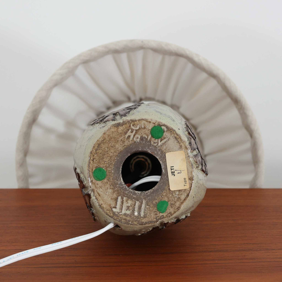 デンマークのJeti Keramikによる、Chris Haslev（クリス・ハスレフ）デザインのビンテージテーブルランプ。陶磁器製のベースは、丸みのある胴部から首元に向かって緩やかに広がる個性的なフォルムが特徴です。上部には素焼きのようなざらりとした質感を残し、下部には淡いグレージュの釉薬と、花を思わせる立体的な装飾が施されています。ブラウンとの落ち着いた色彩の対比が美しく、手仕事ならではの素朴な温かみと豊かな表情が感じられます。プリーツを施したリネンシェードは、点灯すると柔らかな光を広げ、穏やかで心地よい雰囲気を演出します。リビングのサイドテーブルや寝室のチェストなどに取り入れれば、北欧ビンテージらしい趣のある空間を楽しめます。