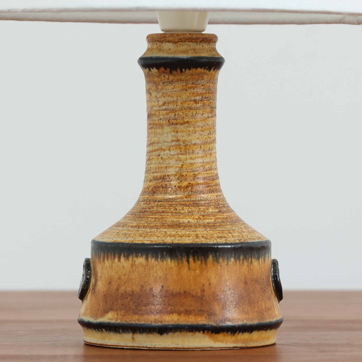デンマークのAxella Keramik（アクセラ）による、Jette Helleroe（イェッテ・ヘレロー）デザインのビンテージテーブルランプ。陶磁器製のベースは、裾に向かって広がる安定感のあるフォルムと、すらりと伸びる首元の美しい曲線が特徴です。温かみのある黄褐色の釉薬に、深いブラウンのラインがアクセントを添え、表面に刻まれた繊細な横筋模様が素朴で味わい深い表情を生み出しています。正面には車輪や花を思わせる円形の装飾が施され、手仕事ならではの個性とクラフト感が漂います。シンプルなリネンシェードは光を柔らかく透過させ、穏やかで温もりのある空間を演出します。リビングのサイドボードや寝室のベッドサイドなどに取り入れれば、北欧ビンテージらしい落ち着いた雰囲気を楽しめます。