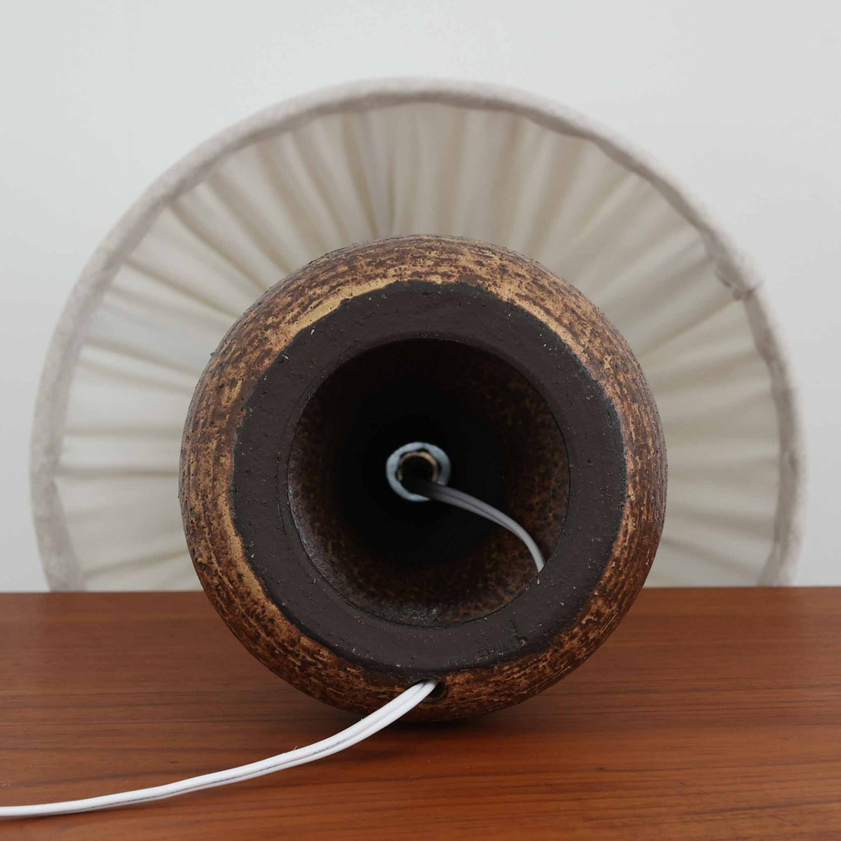 デンマーク製のビンテージテーブルランプ。陶磁器製のベースは、ふっくらと丸みを帯びた下部と、中央が緩やかにくびれた細長い首元を組み合わせた、彫刻的なフォルムが印象的です。深みのあるダークブラウンを基調に、下部には褐色の濃淡が複雑に重なり、ざらりとした質感や釉薬の表情が焼き物ならではの素朴な魅力を引き立てています。落ち着いた色合いながらも存在感があり、北欧ビンテージらしい温もりと力強さを感じさせる佇まいです。細かなプリーツを施したリネンシェードは、点灯すると柔らかな光を広げ、穏やかで心地よい雰囲気を演出します。リビングのサイドボードや寝室のベッドサイドなどにおすすめの一品です。