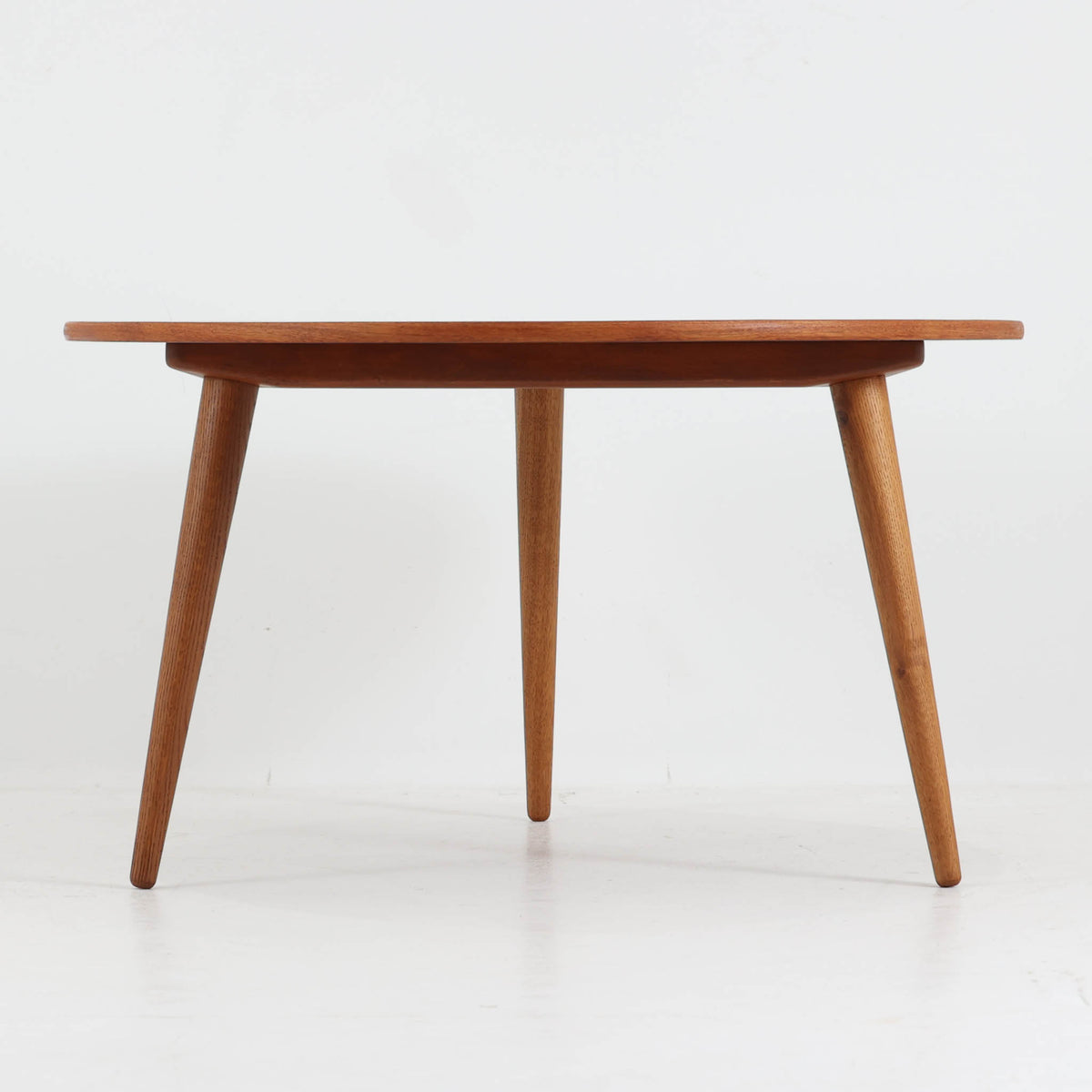 Hans J. Wegnerがデザインし、Andreas Tuckが製造を手掛けたAT8。チーク材の円形天板とオーク材の脚部を組み合わせ、異なる木材の表情を巧みに生かした一台です。直径98cmの伸びやかな天板を、外側へ緩やかに開く4本のテーパードレッグが軽快に支えるシンプルな構成で、素材とプロポーションの美しさが際立ちます。ソファを囲むリビングの主役として取り入れやすく、ウェグナーらしい端正な造形と年月を重ねた木部の風合いを味わえるビンテージ品です。