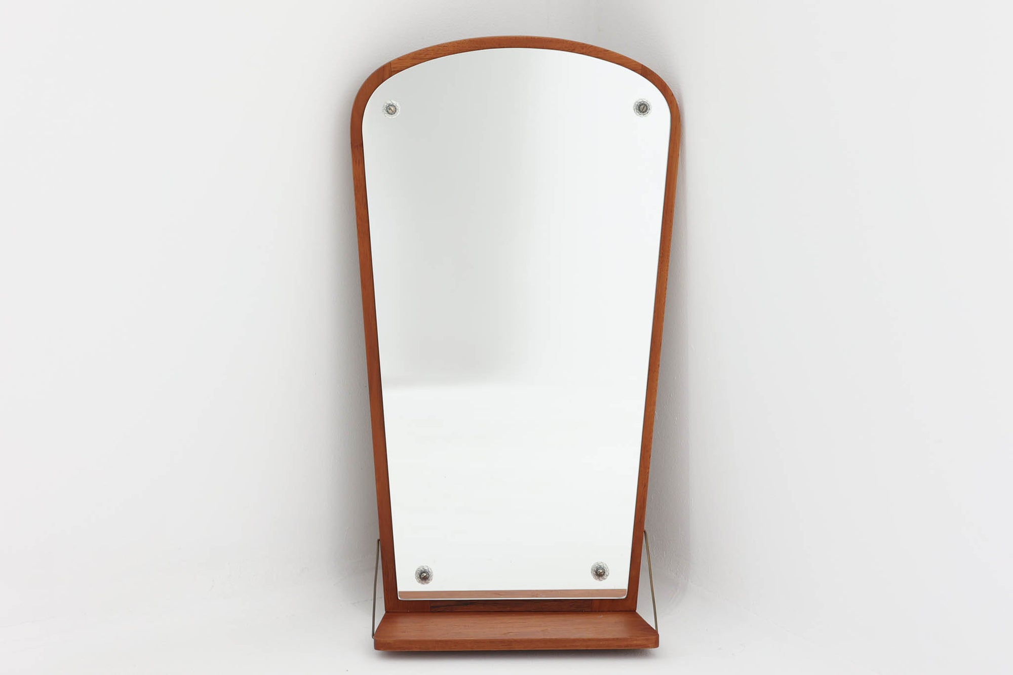 チーク材の美しい木目が魅力の壁掛けミラーは、下部に小さな棚が付いた実用性の高いデザインです。フレームは直線的でありながら角に柔らかな丸みを持たせており、北欧ビンテージ家具ならではの繊細な職人技が感じられます。コンパクトながら存在感があり、玄関先でのちょっとした小物置きや身だしなみチェックに便利です。壁掛け用としても、軽く立て掛けて飾ることもでき、空間を品良く演出してくれます。