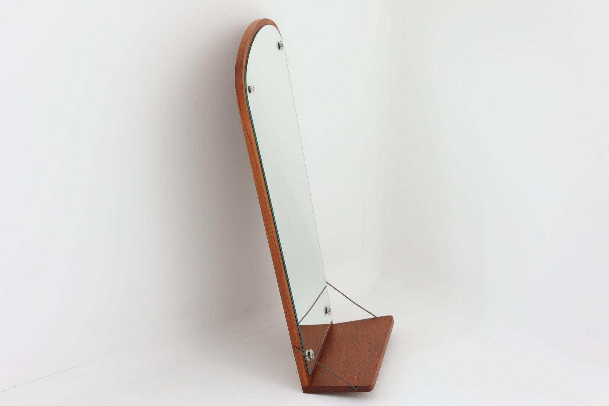 チーク材の美しい木目が魅力の壁掛けミラーは、下部に小さな棚が付いた実用性の高いデザインです。フレームは直線的でありながら角に柔らかな丸みを持たせており、北欧ビンテージ家具ならではの繊細な職人技が感じられます。コンパクトながら存在感があり、玄関先でのちょっとした小物置きや身だしなみチェックに便利です。壁掛け用としても、軽く立て掛けて飾ることもでき、空間を品良く演出してくれます。