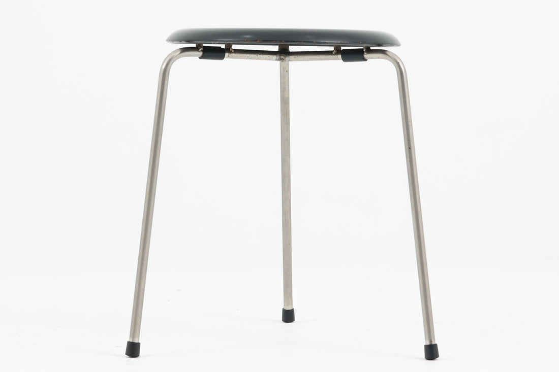 1954年、デンマークのデザイナー、Arne Jacobsen（アルネ・ヤコブセン）によって発表された3本脚のスツール、「DOT（ドット）」。このスツールは、ヤコブセンが手がけた「ルードブレ市庁舎」のロビーで待合席として使用されていました。1970年には4本脚のバージョンも登場しましたが、3本脚のモデルは同年に廃盤となり、現在はビンテージ品のみで入手可能です。オフィスや自宅での日常的な使用はもちろん、予備の椅子としても便利なこのスツールは、シンプルなデザインが魅力です。座面の中央がわずかにくぼんでおり、座った際にフィット感を生み出します。シンプルながらもヤコブセンのこだわりが感じられ、また、単なるスツールとしてだけでなく、ソファの横に置いて本や小物を置くサイドテーブルとしても活用できます。座面裏には薄っすらとFritz Hansen社の刻印がございます。座面に真鍮が施された希少なモデルです。