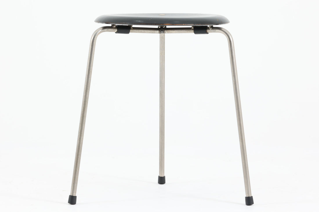 1954年、デンマークのデザイナー、Arne Jacobsen（アルネ・ヤコブセン）によって発表された3本脚のスツール、「DOT（ドット）」。このスツールは、ヤコブセンが手がけた「ルードブレ市庁舎」のロビーで待合席として使用されていました。1970年には4本脚のバージョンも登場しましたが、3本脚のモデルは同年に廃盤となり、現在はビンテージ品のみで入手可能です。オフィスや自宅での日常的な使用はもちろん、予備の椅子としても便利なこのスツールは、シンプルなデザインが魅力です。座面の中央がわずかにくぼんでおり、座った際にフィット感を生み出します。シンプルながらもヤコブセンのこだわりが感じられ、また、単なるスツールとしてだけでなく、ソファの横に置いて本や小物を置くサイドテーブルとしても活用できます。座面裏には薄っすらとFritz Hansen社の刻印がございます。