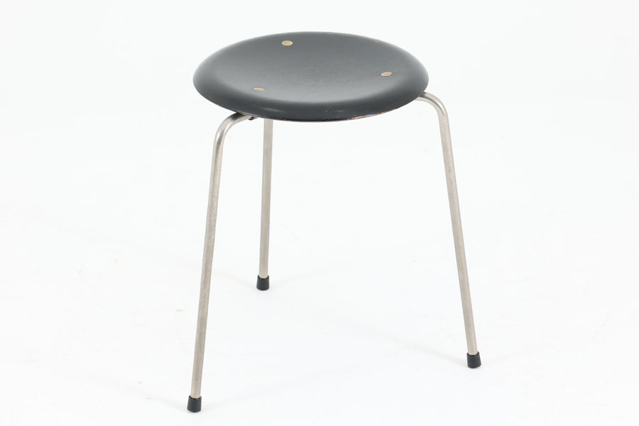 1954年、デンマークのデザイナー、Arne Jacobsen（アルネ・ヤコブセン）によって発表された3本脚のスツール、「DOT（ドット）」。このスツールは、ヤコブセンが手がけた「ルードブレ市庁舎」のロビーで待合席として使用されていました。1970年には4本脚のバージョンも登場しましたが、3本脚のモデルは同年に廃盤となり、現在はビンテージ品のみで入手可能です。オフィスや自宅での日常的な使用はもちろん、予備の椅子としても便利なこのスツールは、シンプルなデザインが魅力です。座面の中央がわずかにくぼんでおり、座った際にフィット感を生み出します。シンプルながらもヤコブセンのこだわりが感じられ、また、単なるスツールとしてだけでなく、ソファの横に置いて本や小物を置くサイドテーブルとしても活用できます。座面裏には薄っすらとFritz Hansen社の刻印がございます。