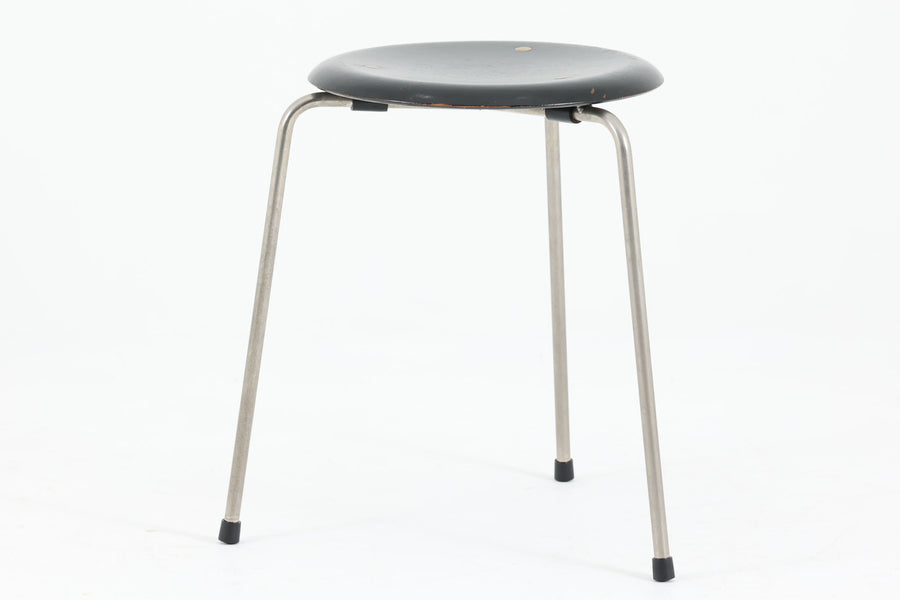 1954年、デンマークのデザイナー、Arne Jacobsen（アルネ・ヤコブセン）によって発表された3本脚のスツール、「DOT（ドット）」。このスツールは、ヤコブセンが手がけた「ルードブレ市庁舎」のロビーで待合席として使用されていました。1970年には4本脚のバージョンも登場しましたが、3本脚のモデルは同年に廃盤となり、現在はビンテージ品のみで入手可能です。オフィスや自宅での日常的な使用はもちろん、予備の椅子としても便利なこのスツールは、シンプルなデザインが魅力です。座面の中央がわずかにくぼんでおり、座った際にフィット感を生み出します。シンプルながらもヤコブセンのこだわりが感じられ、また、単なるスツールとしてだけでなく、ソファの横に置いて本や小物を置くサイドテーブルとしても活用できます。座面裏には薄っすらとFritz Hansen社の刻印がございます。