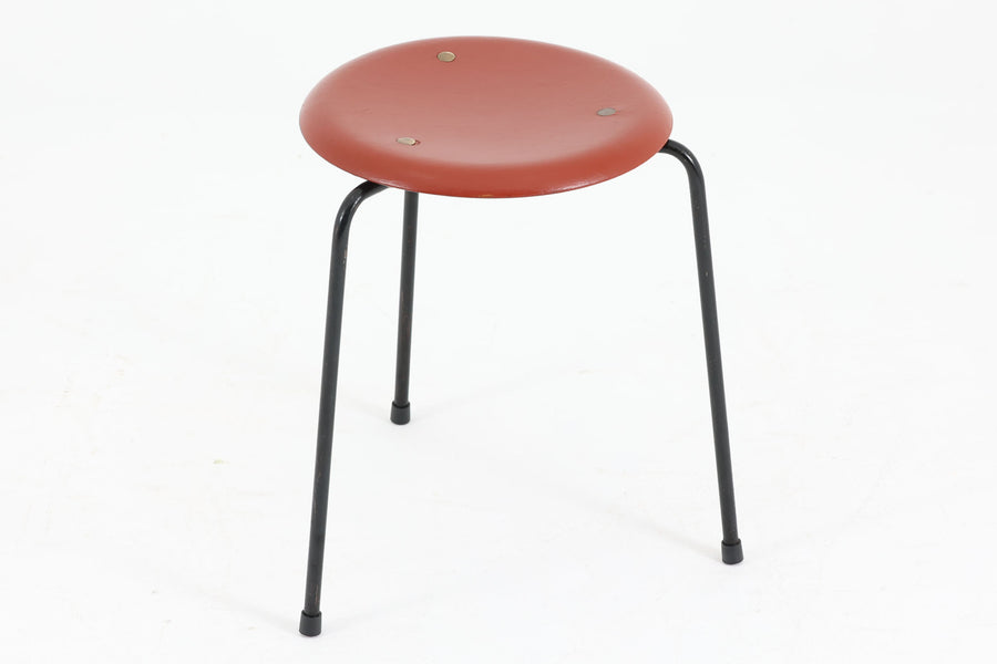 1954年、デンマークのデザイナー、Arne Jacobsen（アルネ・ヤコブセン）によって発表された3本脚のスツール、「DOT（ドット）」。このスツールは、ヤコブセンが手がけた「ルードブレ市庁舎」のロビーで待合席として使用されていました。1970年には4本脚のバージョンも登場しましたが、3本脚のモデルは同年に廃盤となり、現在はビンテージ品のみで入手可能です。オフィスや自宅での日常的な使用はもちろん、予備の椅子としても便利なこのスツールは、シンプルなデザインが魅力です。座面の中央がわずかにくぼんでおり、座った際にフィット感を生み出します。シンプルながらもヤコブセンのこだわりが感じられ、また、単なるスツールとしてだけでなく、ソファの横に置いて本や小物を置くサイドテーブルとしても活用できます。前オーナーによってリペイントされています。座面裏には薄っすらとFritz Hansen社の刻印がございます。