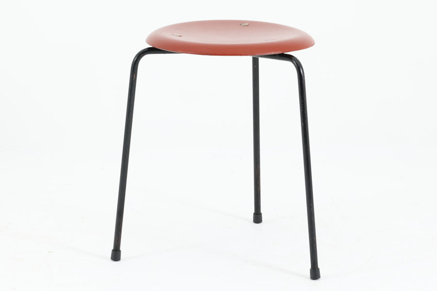 1954年、デンマークのデザイナー、Arne Jacobsen（アルネ・ヤコブセン）によって発表された3本脚のスツール、「DOT（ドット）」。このスツールは、ヤコブセンが手がけた「ルードブレ市庁舎」のロビーで待合席として使用されていました。1970年には4本脚のバージョンも登場しましたが、3本脚のモデルは同年に廃盤となり、現在はビンテージ品のみで入手可能です。オフィスや自宅での日常的な使用はもちろん、予備の椅子としても便利なこのスツールは、シンプルなデザインが魅力です。座面の中央がわずかにくぼんでおり、座った際にフィット感を生み出します。シンプルながらもヤコブセンのこだわりが感じられ、また、単なるスツールとしてだけでなく、ソファの横に置いて本や小物を置くサイドテーブルとしても活用できます。前オーナーによってリペイントされています。座面裏には薄っすらとFritz Hansen社の刻印がございます。